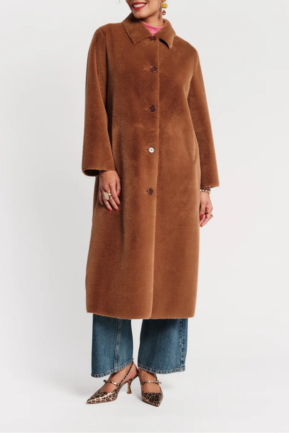 A woman modeling a long brown faux-fur coat with a collar and front buttons, paired with blue jeans and leopard-print heels, against a plain white studio background.