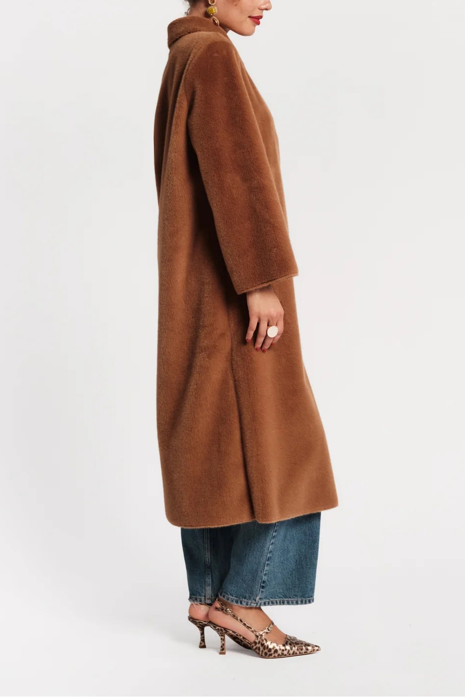A woman modeling a long, brown faux fur coat from the side against a plain white studio background.