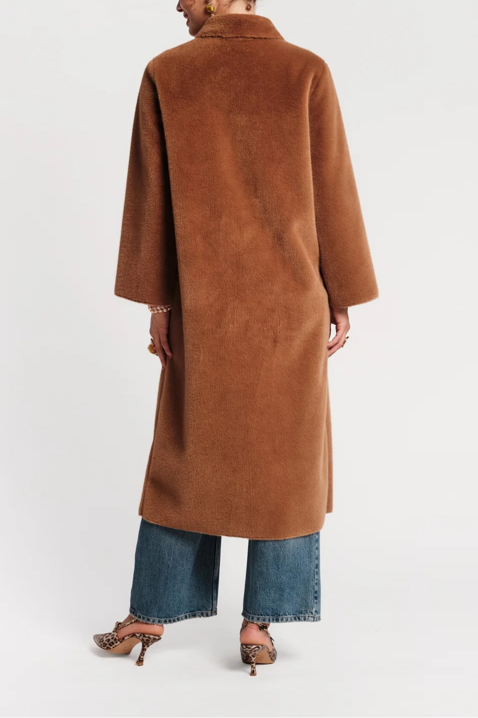A woman shown from the back wearing a long, camel-brown suede or faux-suede coat over blue wide-leg jeans and leopard-print heeled sandals, photographed against a clean white studio background.