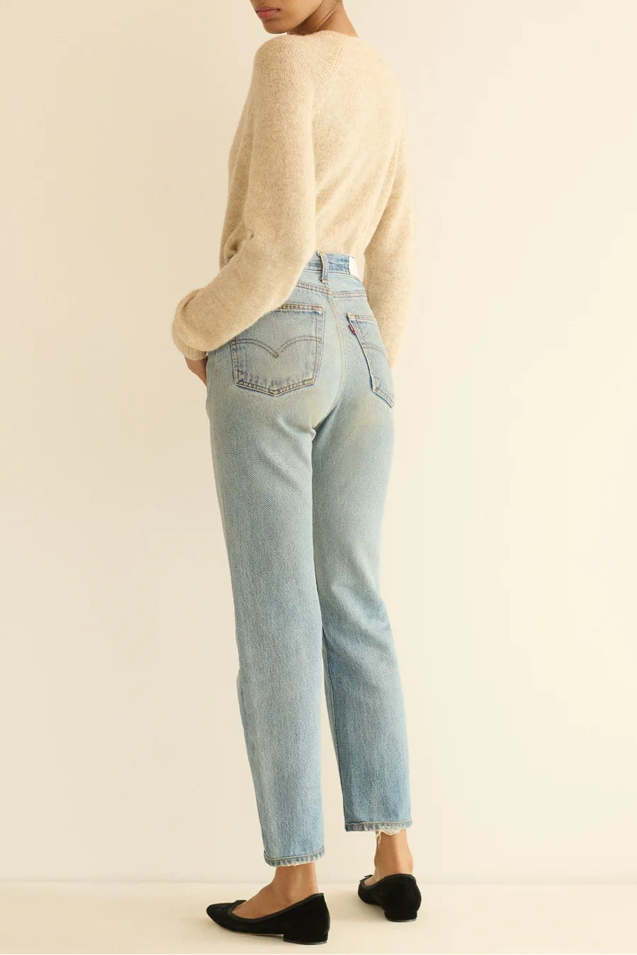 A woman stands in profile wearing a cream knit sweater, light-wash blue jeans, and black flats against a plain off-white studio background.