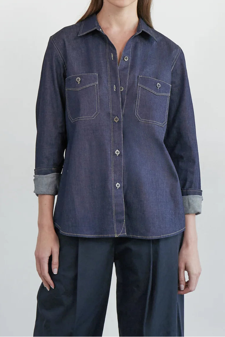 A woman modeling a dark blue denim button-up shirt with two chest pockets and rolled cuffs, paired with dark pants, against a plain light gray studio background.