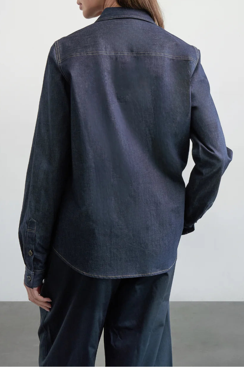 A woman shown from the back wearing a dark indigo denim shirt jacket and matching dark trousers against a plain light gray studio background.