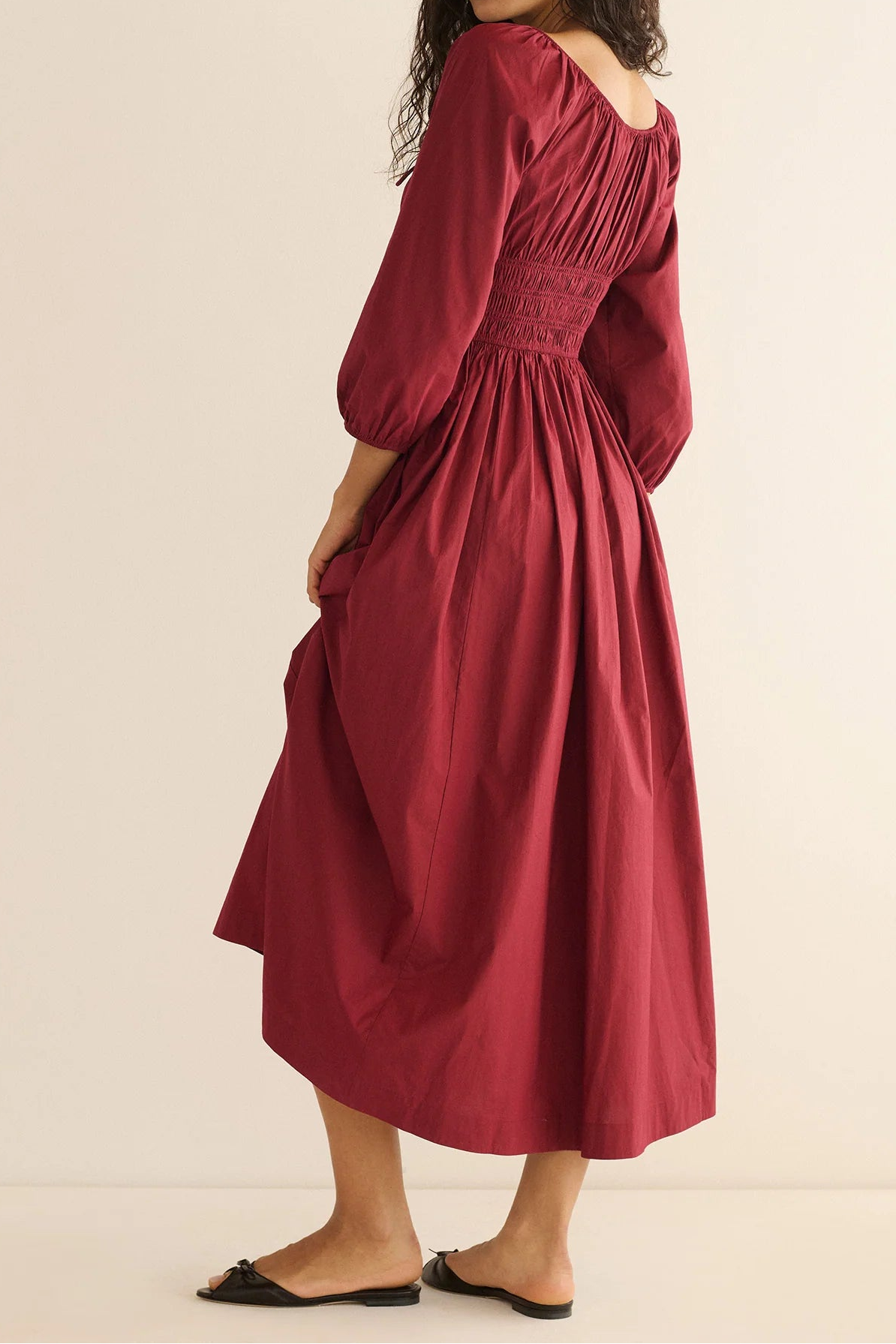 A woman wearing a long-sleeved midi dress in a deep burgundy red, shown from a side-back angle against a plain beige studio background.