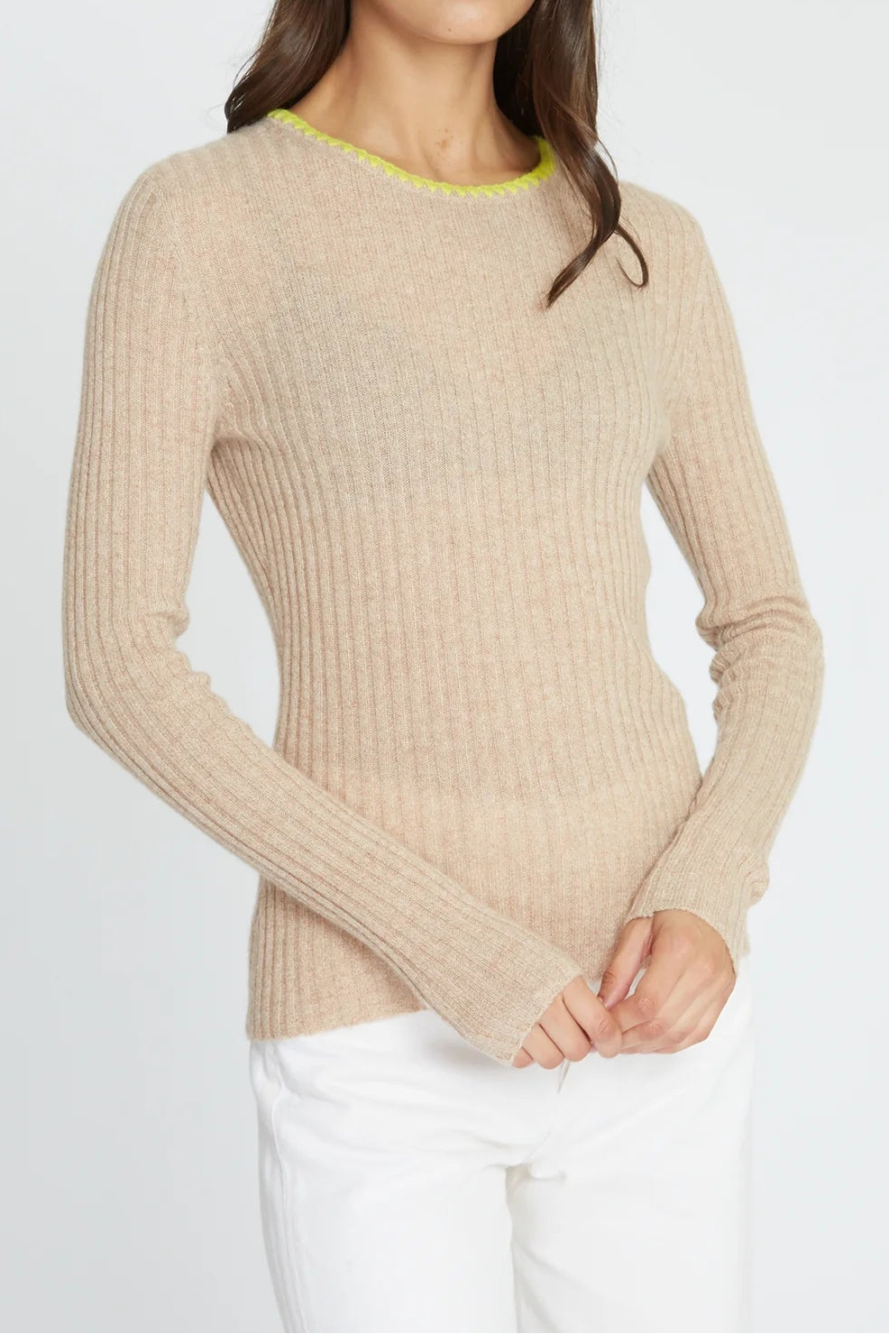 A woman modeling a beige ribbed knit crewneck sweater with a bright yellow-green trim at the collar, paired with white pants, against a light gray studio background.