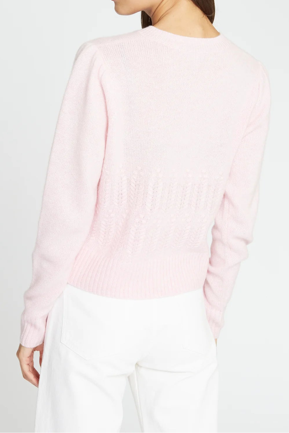 A woman shown from the back wearing a light pink knit sweater and white trousers against a plain light gray studio background.