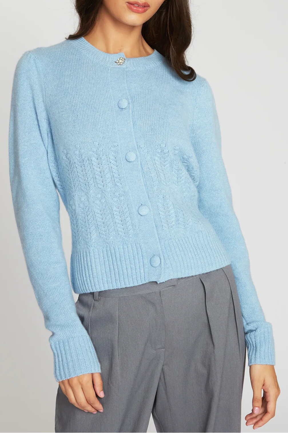 A woman modeling a light blue cropped knit cardigan sweater with front buttons, paired with gray trousers, against a plain light gray studio background.