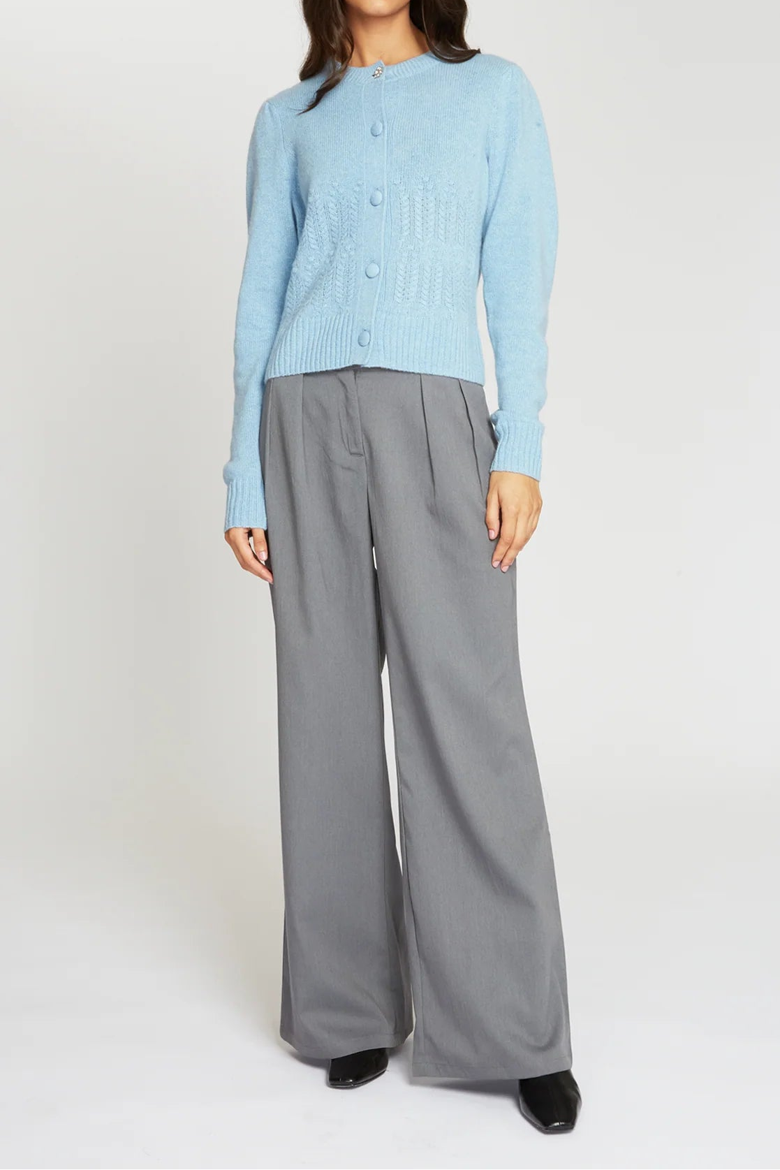A woman modeling a light blue ribbed knit cardigan sweater paired with wide-leg gray trousers and black shoes, standing against a plain light gray studio background.
