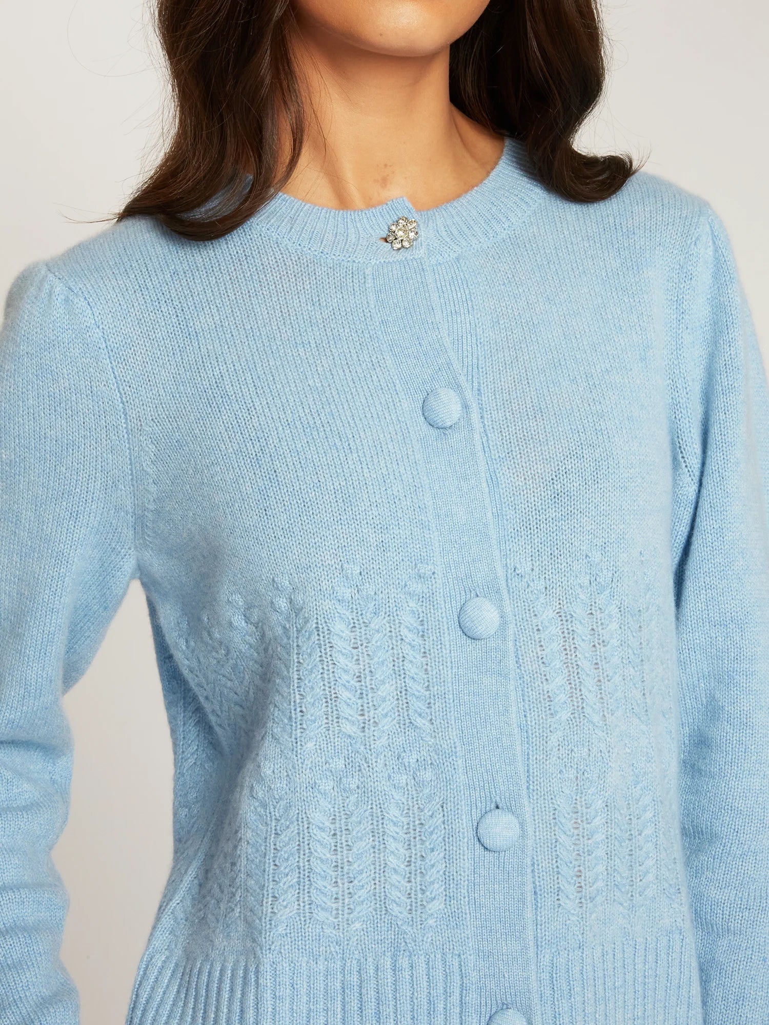 A close-up of a woman wearing a light blue knit cardigan sweater with a decorative silver-toned brooch at the neckline and matching buttons down the front.