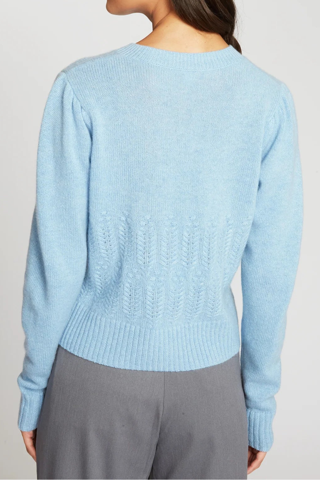 A woman shown from the back wearing a light blue knit sweater and gray pants against a plain light gray studio background.