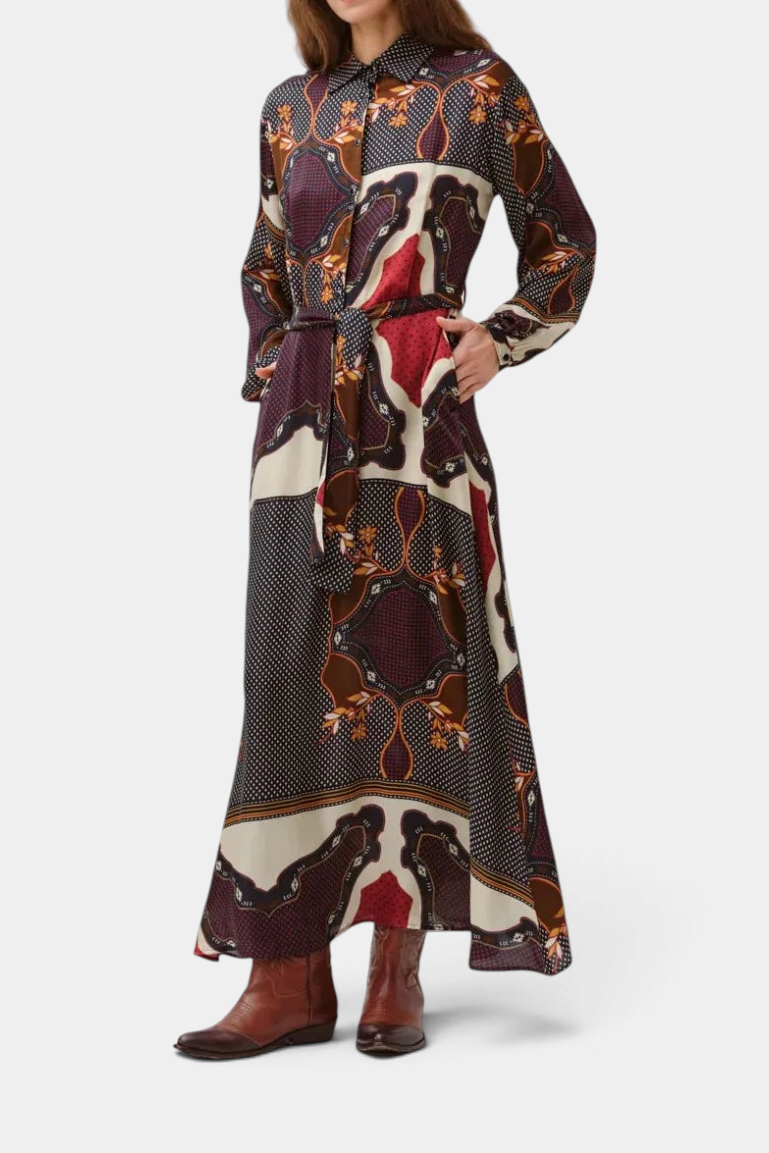 A woman modeling a long-sleeved maxi shirt dress with a complex abstract pattern in dark purple, brown, red, and cream, paired with brown leather boots against a white studio background.