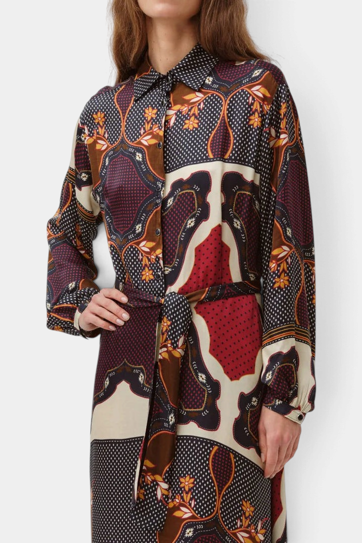 A woman modeling a long-sleeve collared shirt dress with a complex multicolor patchwork print featuring red, brown, beige, and black patterns, set against a plain light gray background.