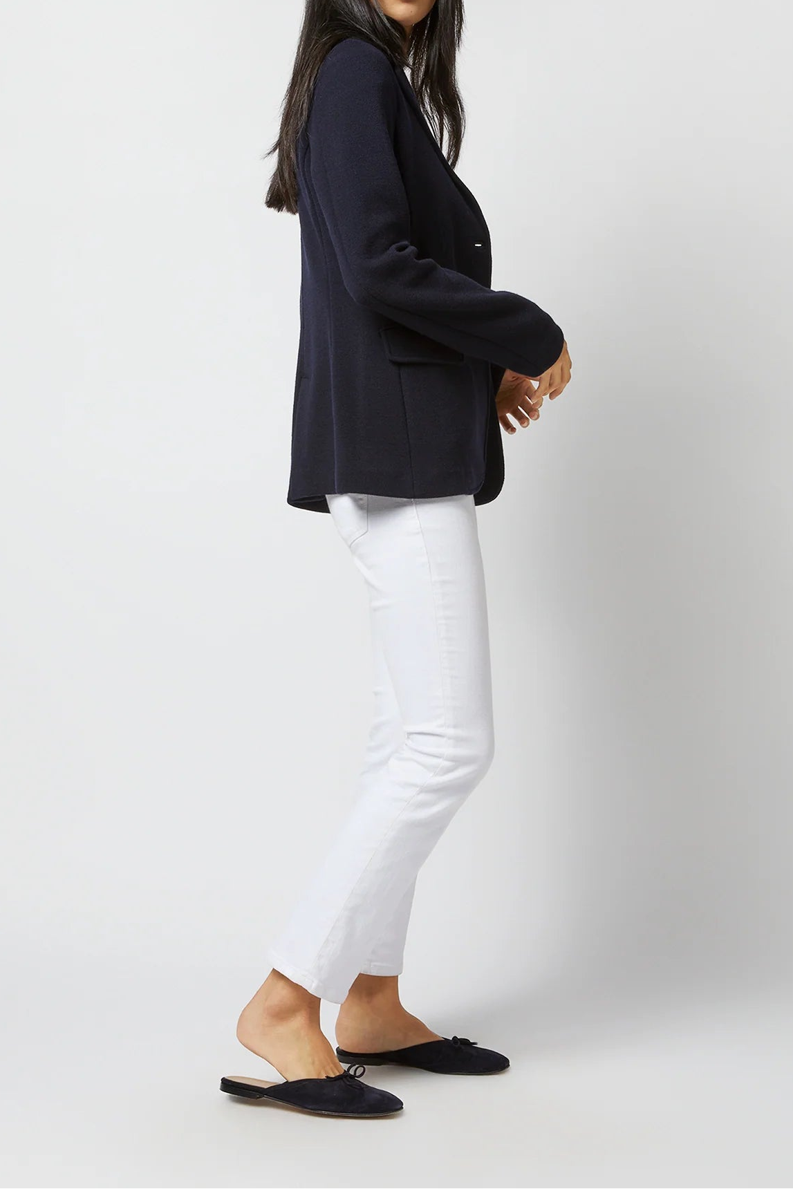 A woman modeling a dark navy blazer, white cropped pants, and black mule flats against a light gray studio background.