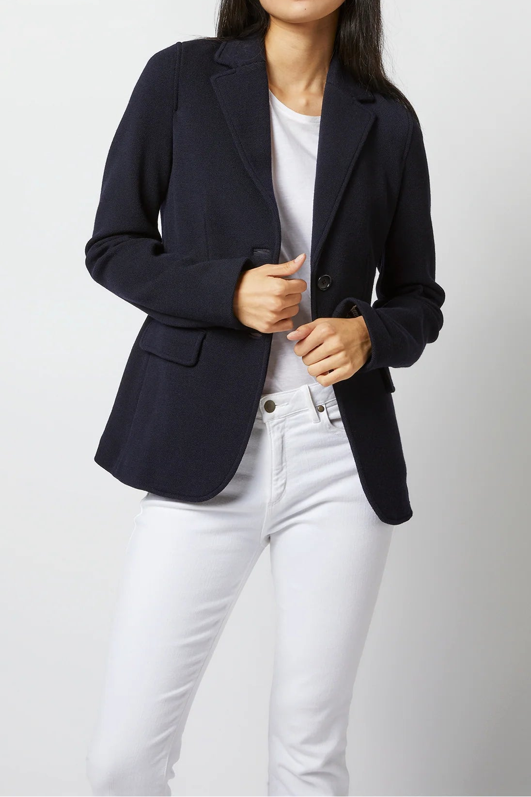 A woman modeling a dark navy blue blazer over a white t-shirt and white jeans, standing against a light gray studio background.