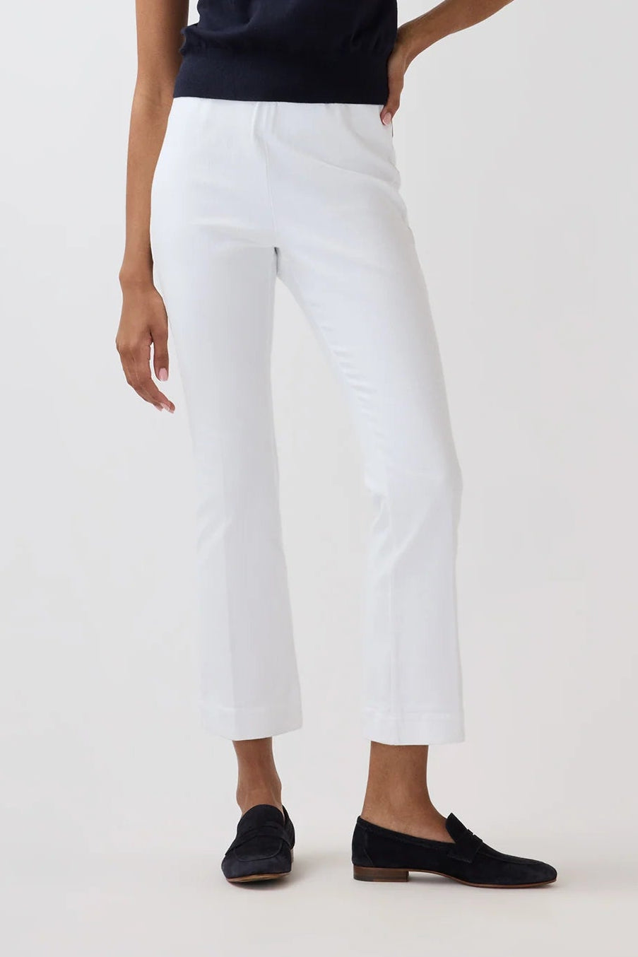 A woman modeling a sleeveless navy top and white cropped pants with black loafers, standing against a plain light gray studio background.