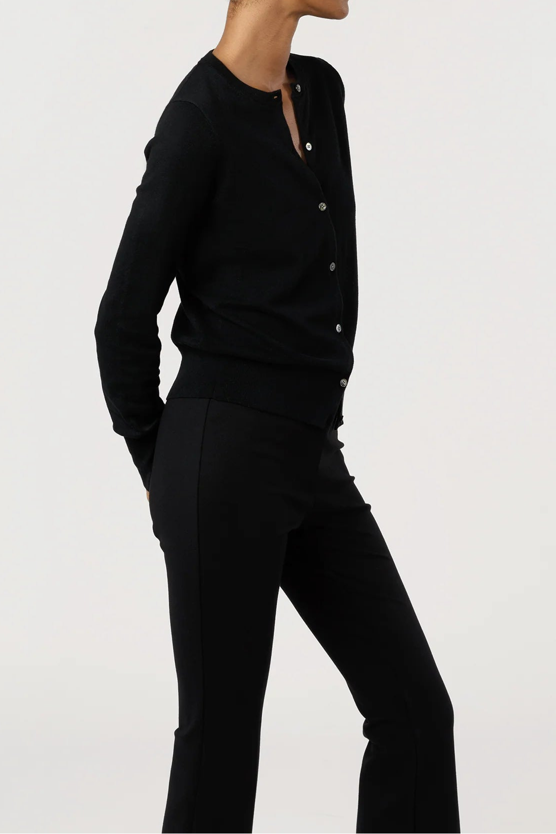 A model in profile wearing a black long-sleeve button-front cardigan and matching black pants, photographed against a clean light gray studio background.