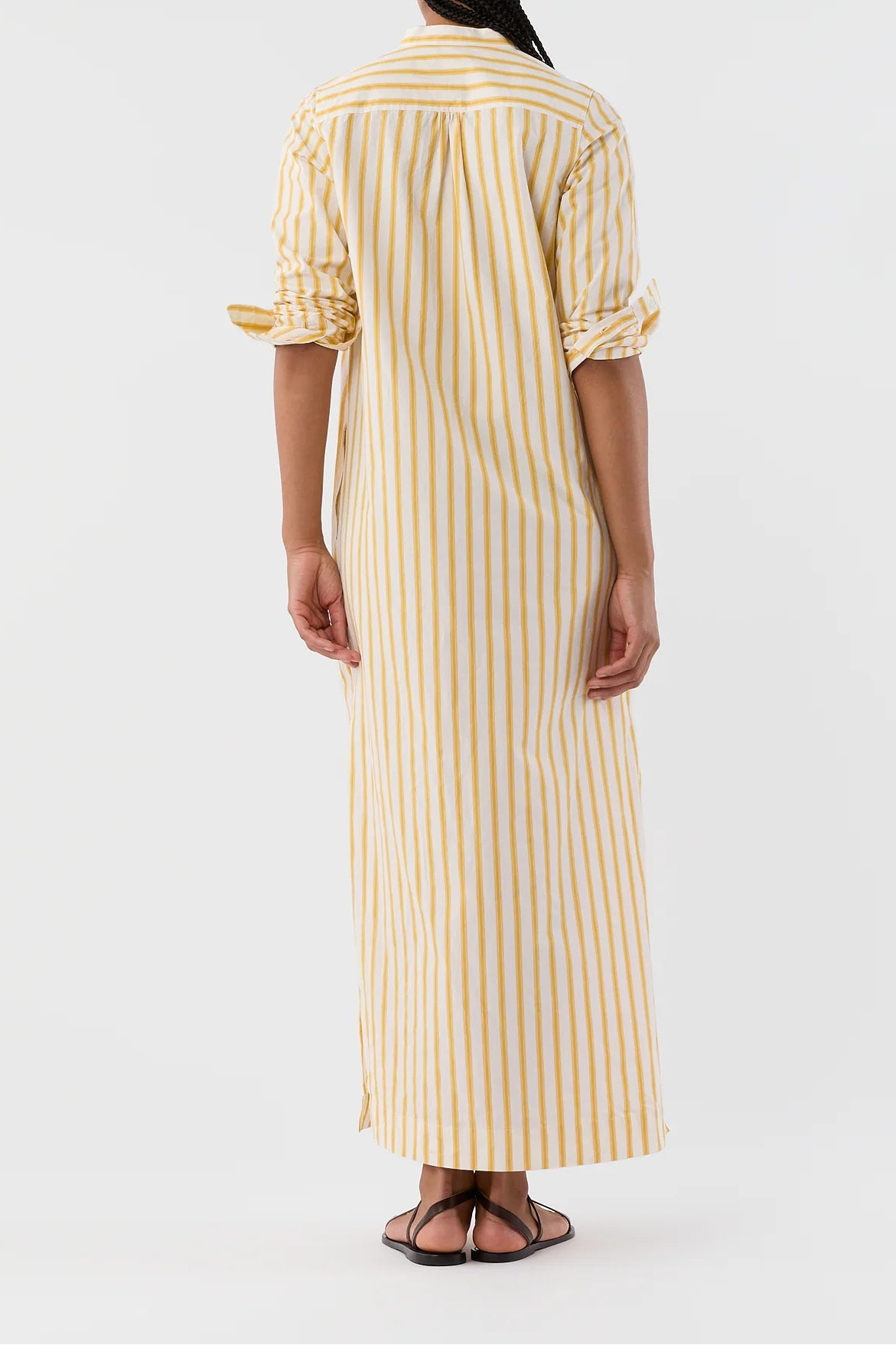 A woman viewed from the back wearing a long yellow and white vertically striped shirt dress with rolled-up sleeves, paired with dark sandals, against a plain light gray studio background.