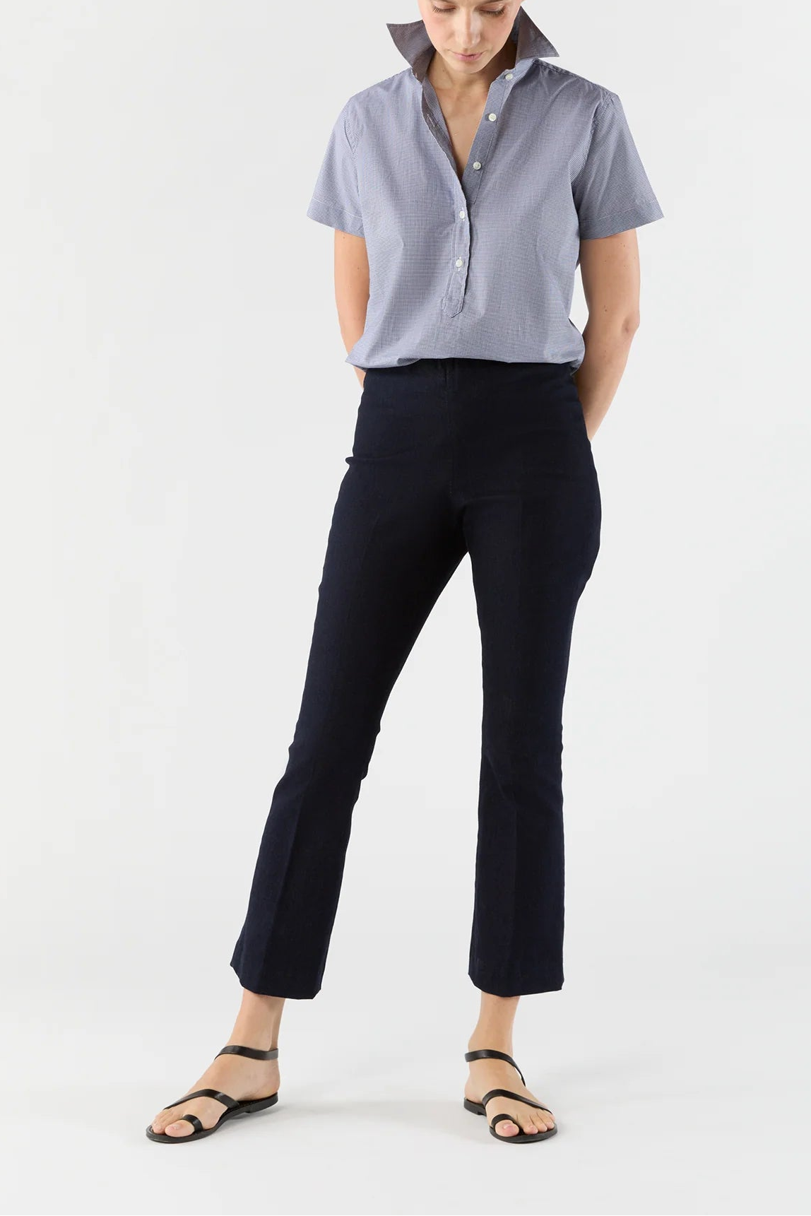 A woman modeling a light blue short-sleeve button-up shirt and dark navy cropped pants with black sandals, photographed against a plain white studio background.