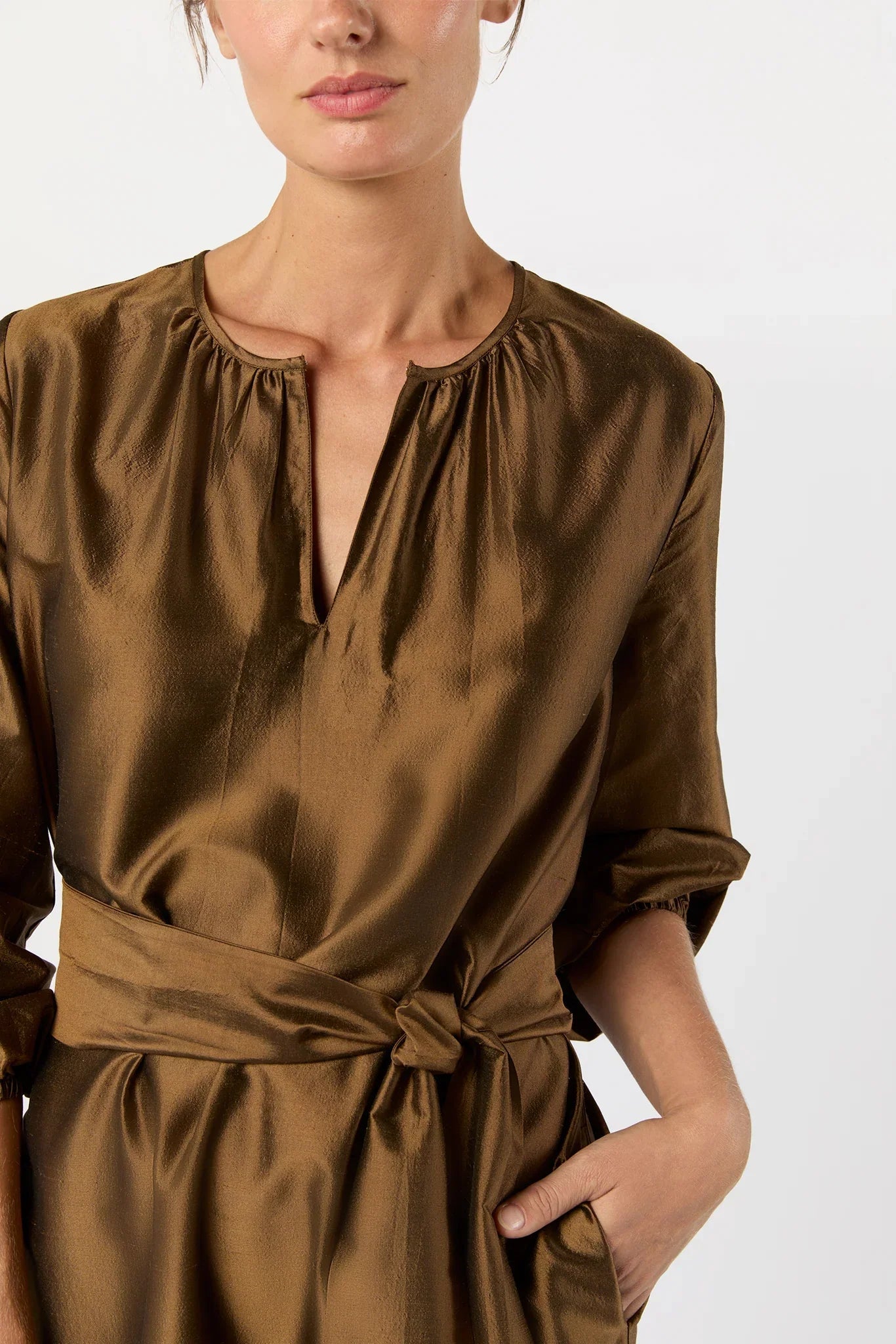 A close-up of a model wearing a brown satin dress with a gathered neckline, tie waist, and elbow-length sleeves against a plain light background.