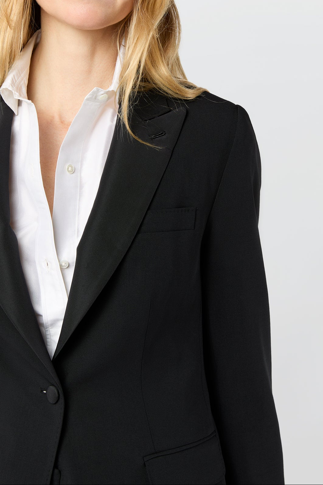 A woman wearing a black blazer over a white collared shirt, photographed against a plain light gray studio background.