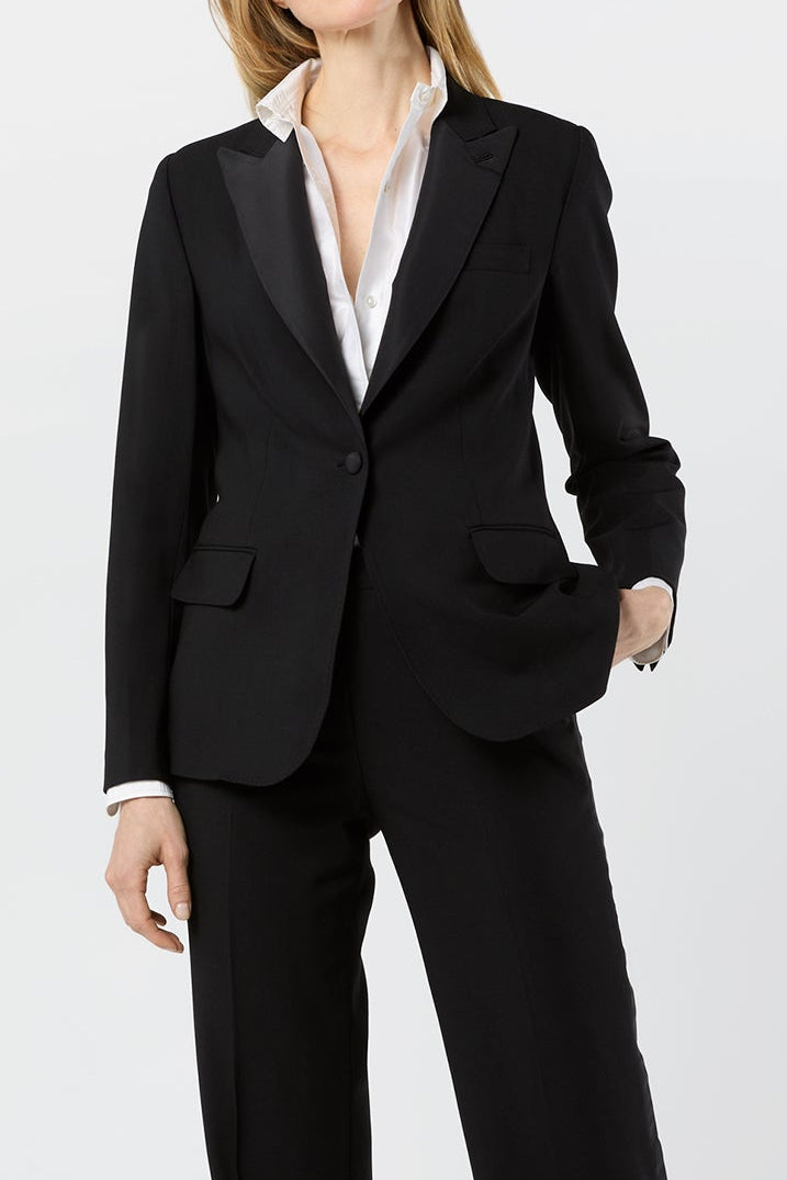A woman modeling a black tailored pantsuit with a single-breasted blazer, wide-leg trousers, and black pointed-toe heels, worn over a white collared shirt against a light gray studio background.