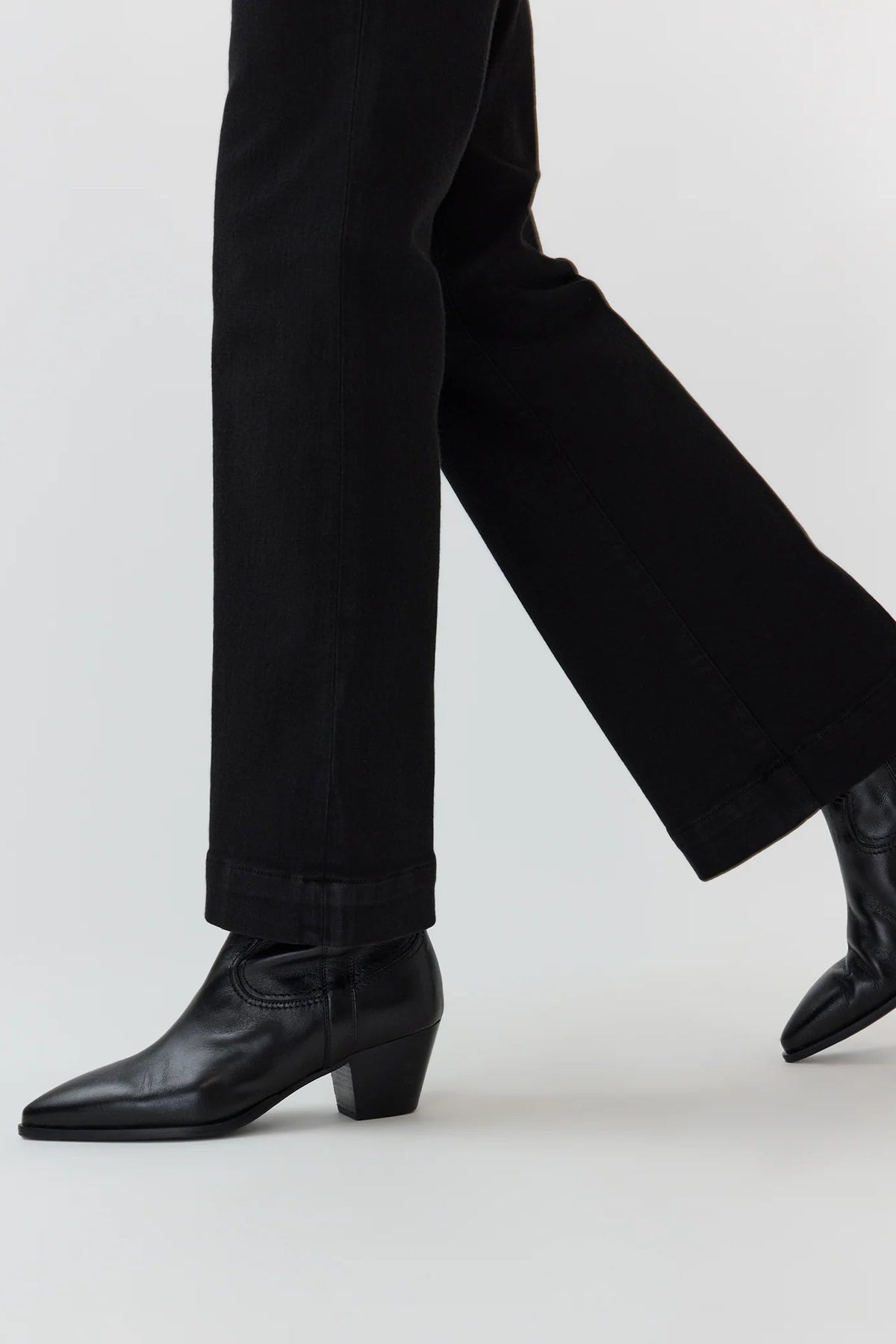 Black leather ankle boots with pointed toes and low block heels, shown on a model wearing black wide-leg jeans against a light gray studio background.