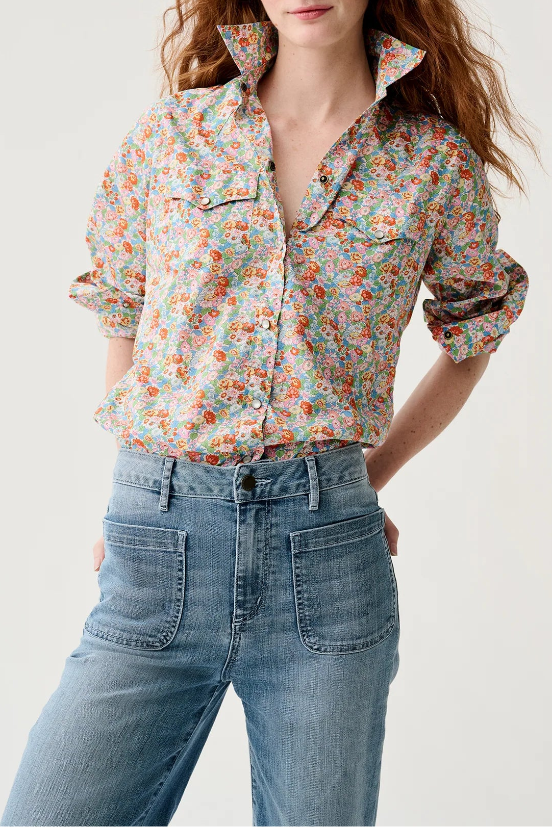 A woman modeling a long-sleeve button-up shirt with a dense multicolor floral print on a light beige base, paired with blue jeans, against a plain light gray background.