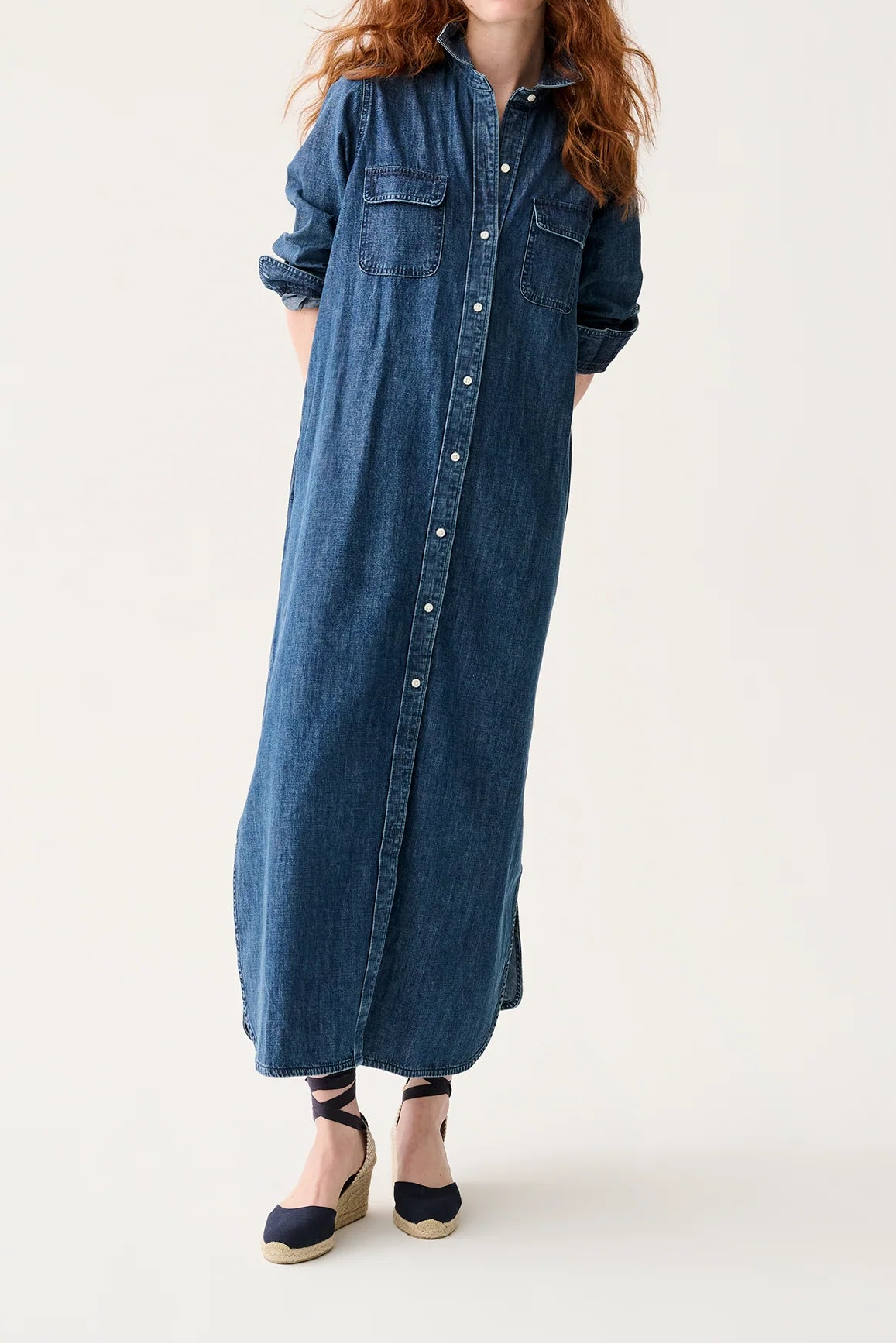 A woman with long red hair wearing a long blue denim shirt dress with rolled-up sleeves and dark wedge sandals, standing against a plain off-white studio background.