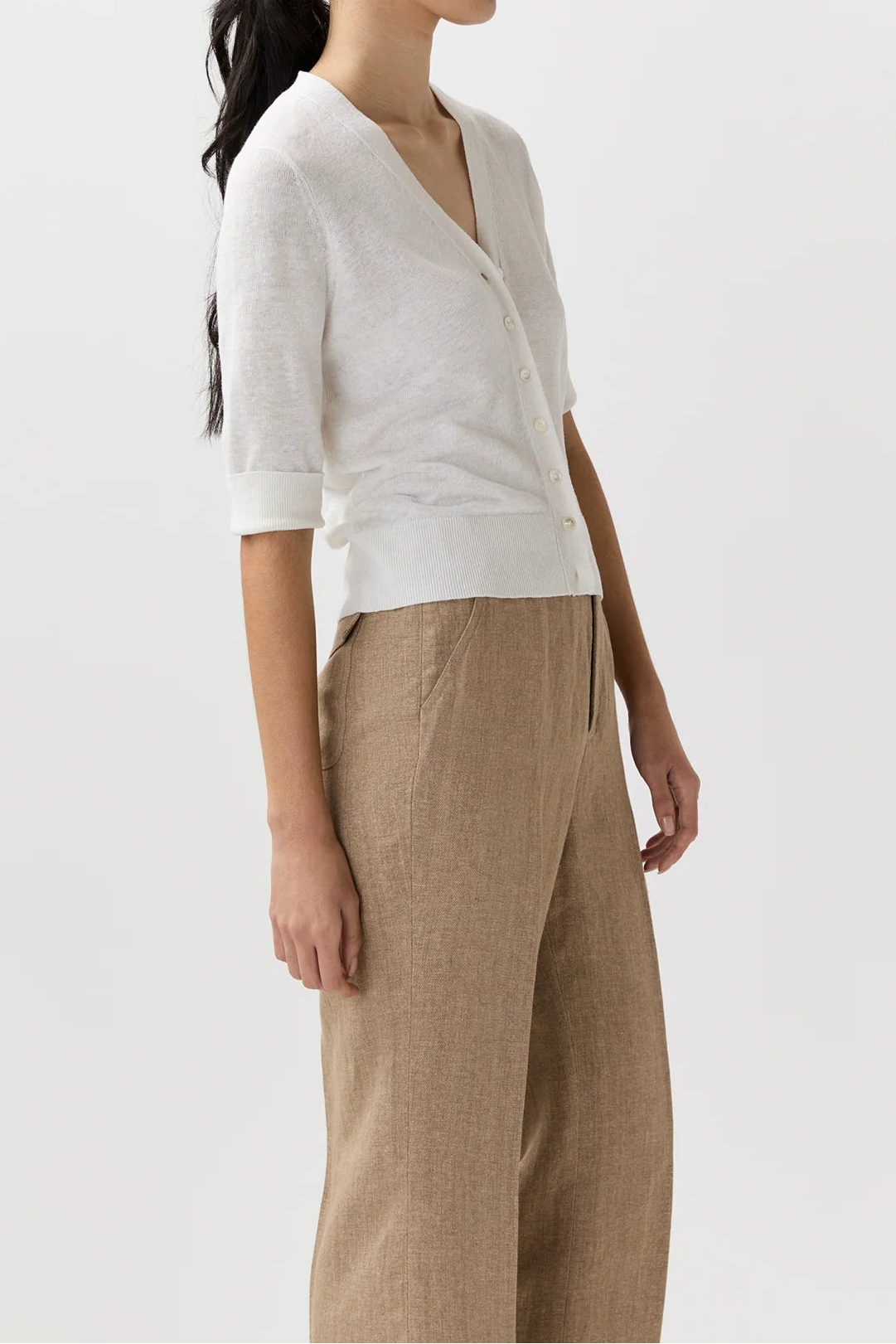 A woman in side profile wearing a white short-sleeve knit cardigan and beige wide-leg trousers, photographed against a clean off-white studio background.