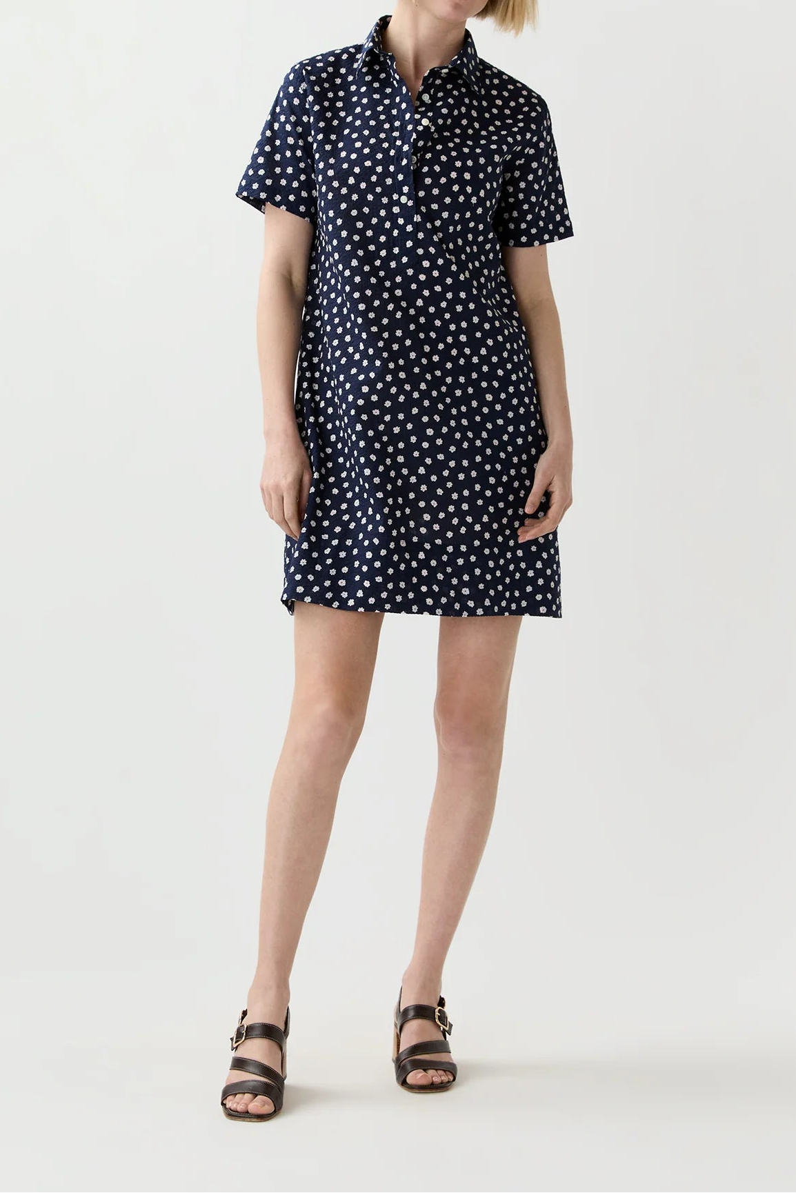 A woman modeling a short-sleeve navy blue shirt dress with a white polka dot pattern, paired with brown double-strap sandals, against a plain light gray studio background.
