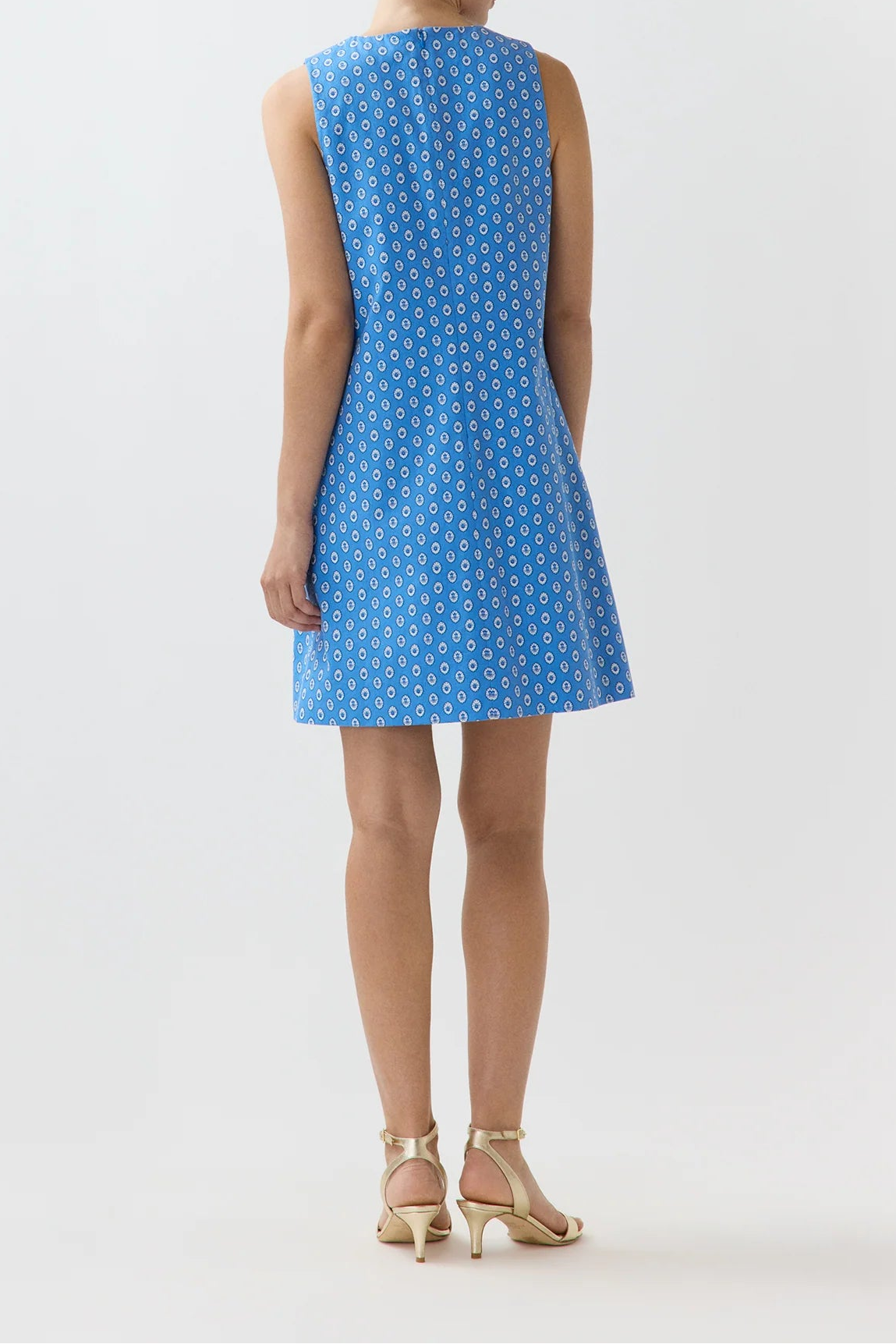 A woman viewed from the back wearing a sleeveless blue shift dress with a white geometric pattern, paired with gold high-heeled sandals, against a plain white studio background.
