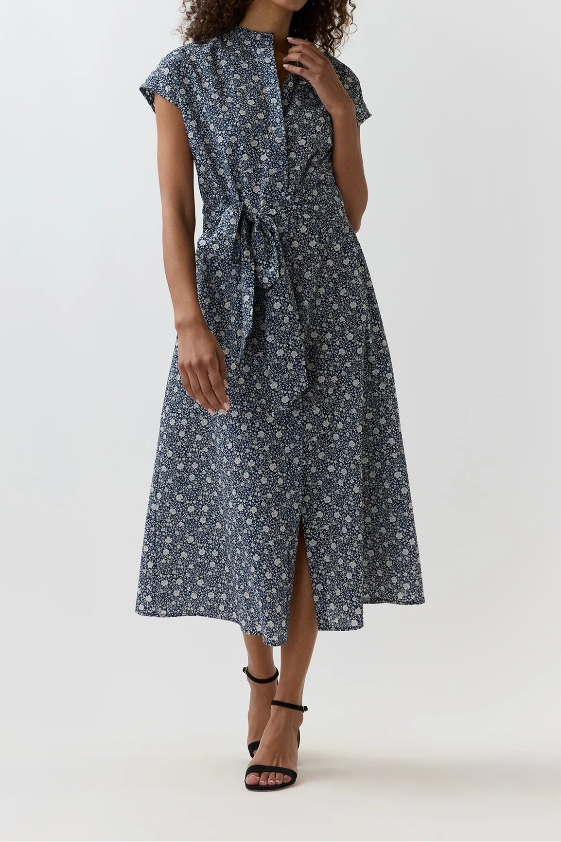 A woman modeling a short-sleeve midi dress with a blue and white floral pattern, featuring a wrap-style front and a self-tie belt at the waist, set against a plain light gray studio background.
