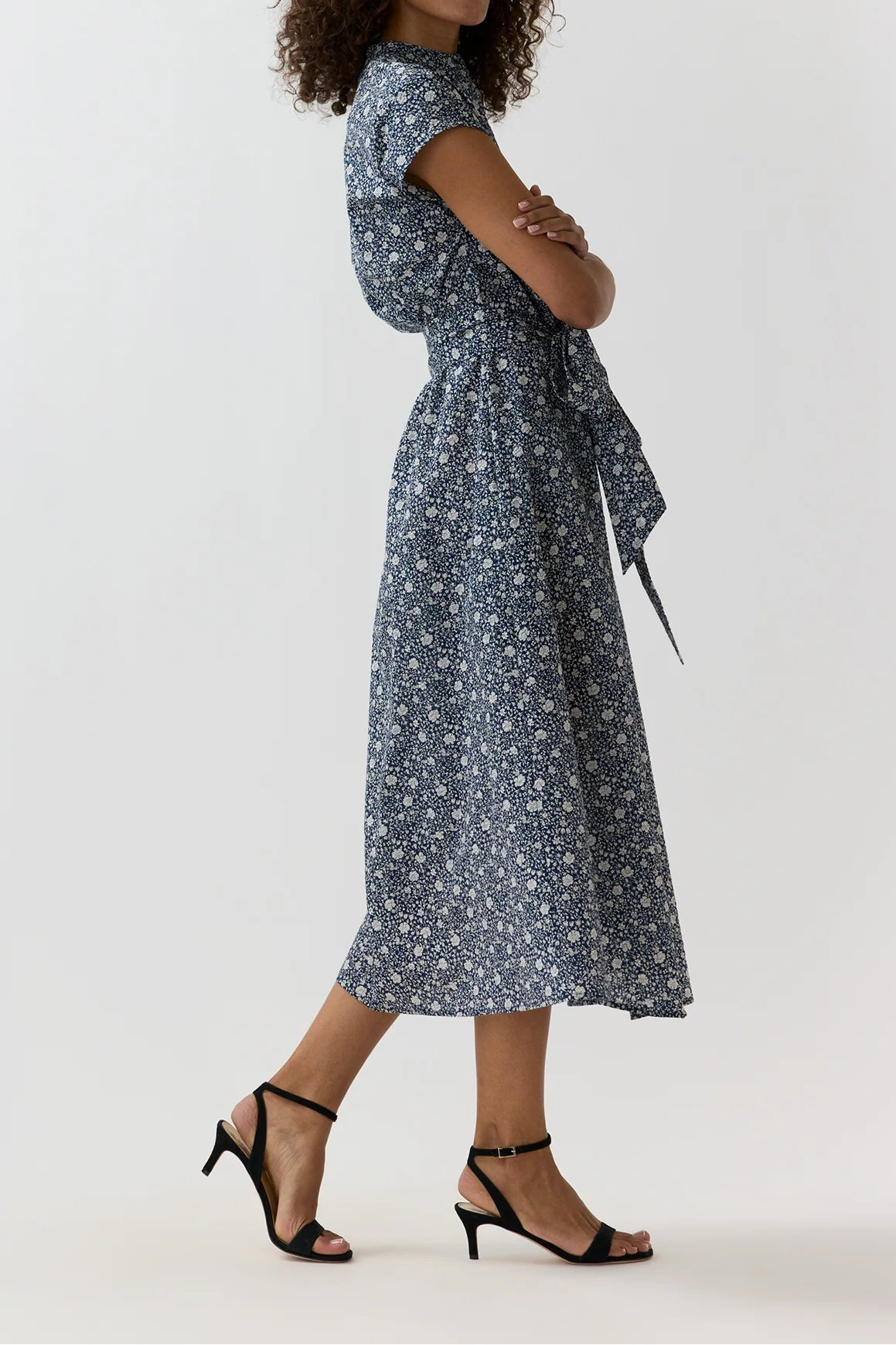 A woman modeling a short-sleeve midi dress with a white floral print on a slate blue-gray background, paired with black strappy high-heeled sandals, against a plain light gray studio background.