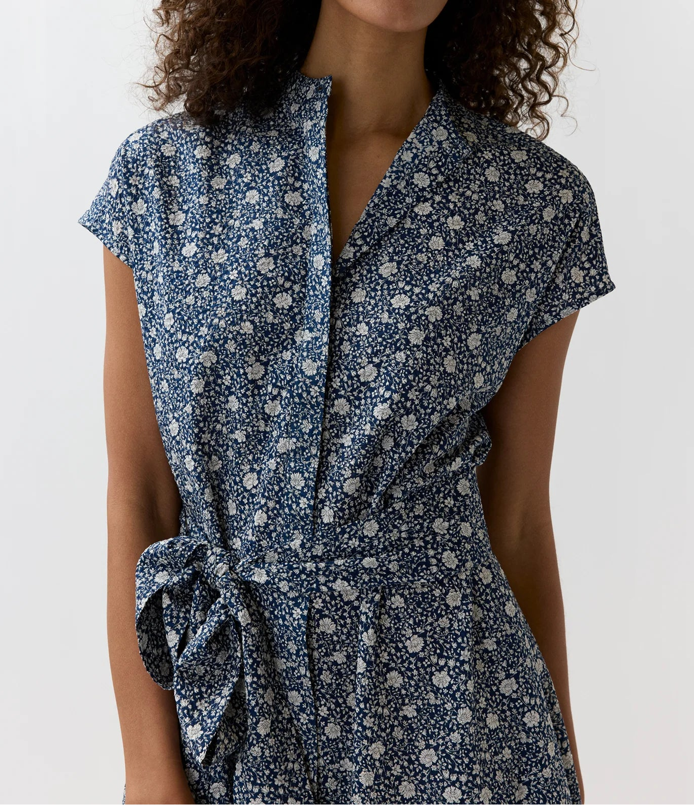 A woman with curly brown hair wearing a navy blue short-sleeve wrap dress with a white floral pattern and a tie waist, photographed against a plain white studio background.