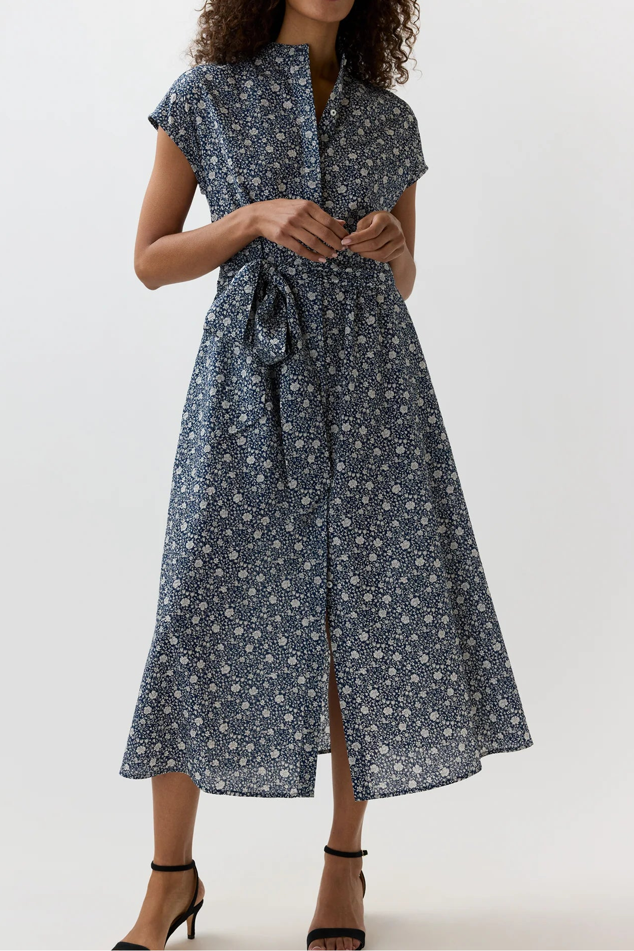 A woman modeling a navy blue midi dress with a white floral print, featuring short sleeves, a high collar, and a self-tie waist belt, set against a plain light gray background.