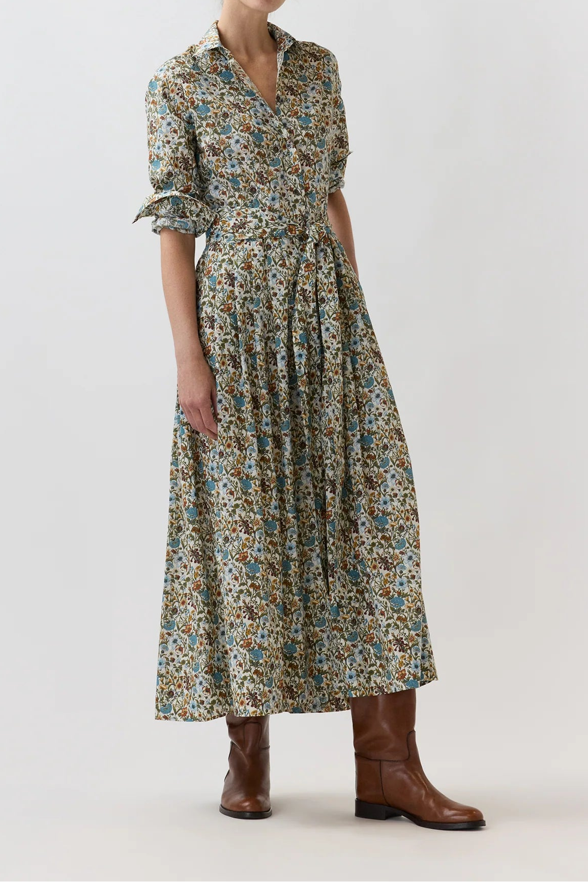 A woman modeling a long-sleeved midi dress with a dense multicolor floral print in blue, green, and yellow tones on a beige base, paired with brown leather boots, against a plain light gray studio background.