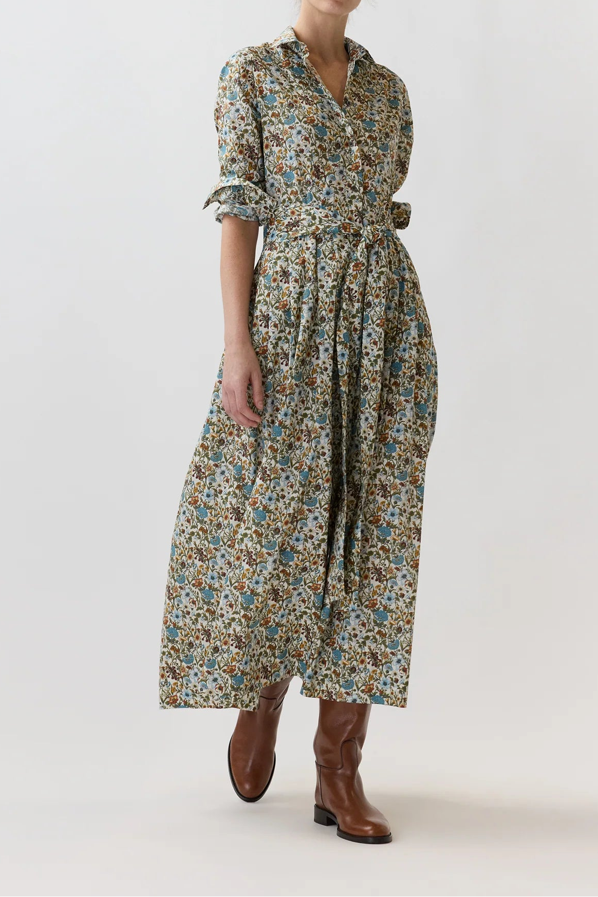 A woman modeling a long-sleeved midi dress with a dense multicolor floral print in blue, green, and yellow on a beige base, paired with brown leather boots, against a plain light gray studio background.
