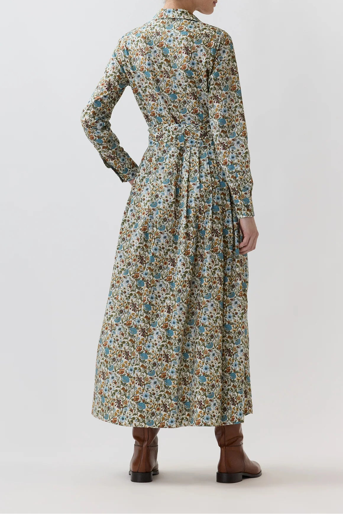 A woman stands with her back to the camera wearing a long-sleeved midi dress with a dense multicolor floral print in blue, green, yellow, and brown on a cream base, paired with brown ankle boots, against a plain light gray studio background.