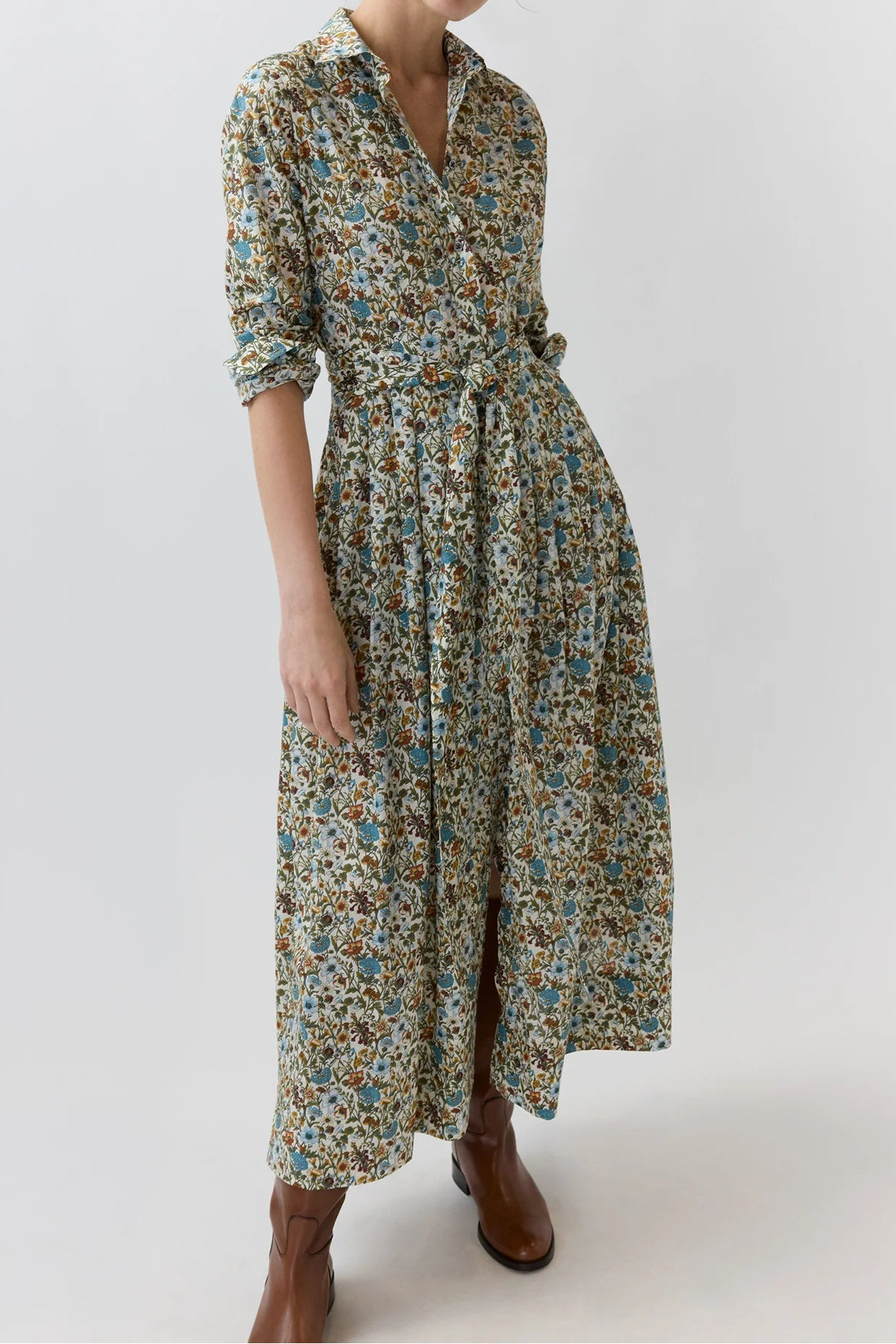 A woman modeling a long-sleeved midi dress with a dense multicolor floral print in blue, green, and brown tones, paired with brown ankle boots, against a plain light gray studio background.