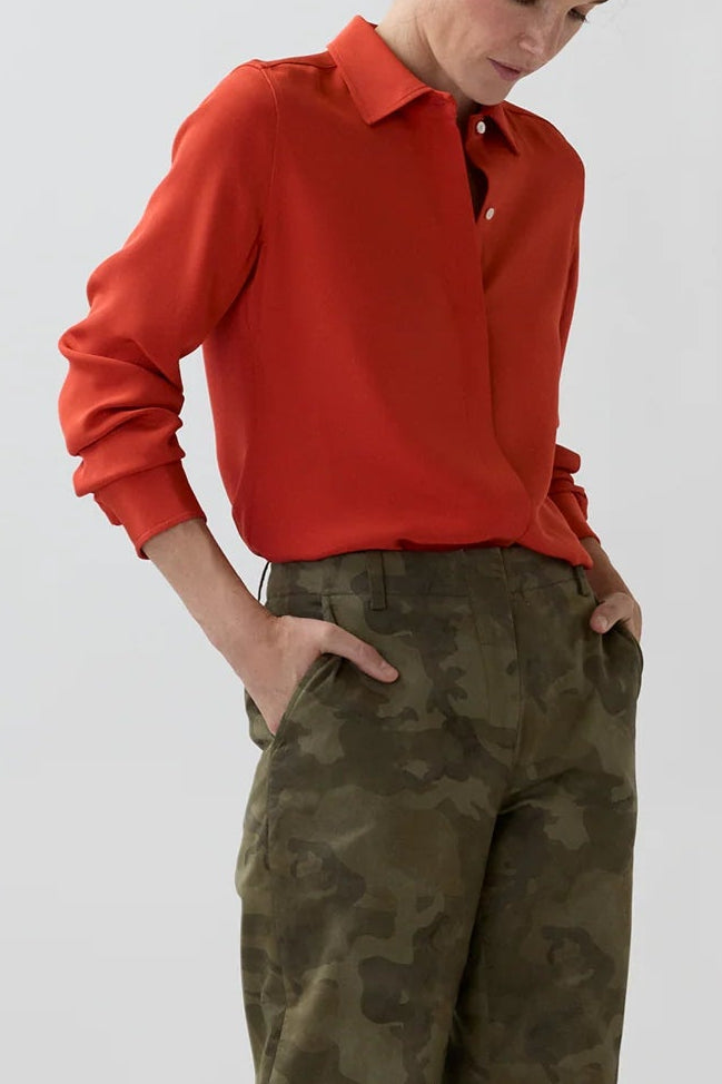 A woman modeling a red long-sleeve collared shirt and olive green camouflage trousers, paired with tan heels, against a plain light gray studio background.