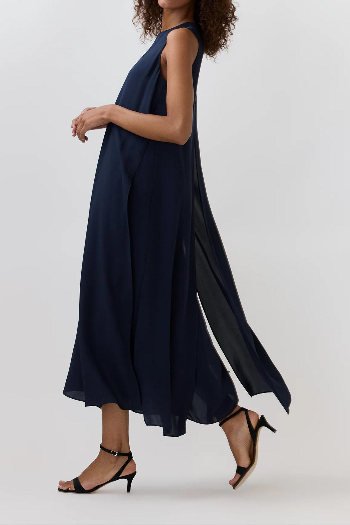 A woman modeling a sleeveless navy blue maxi dress with a flowing, draped design and side slit, paired with black strappy heels, against a light gray studio background.