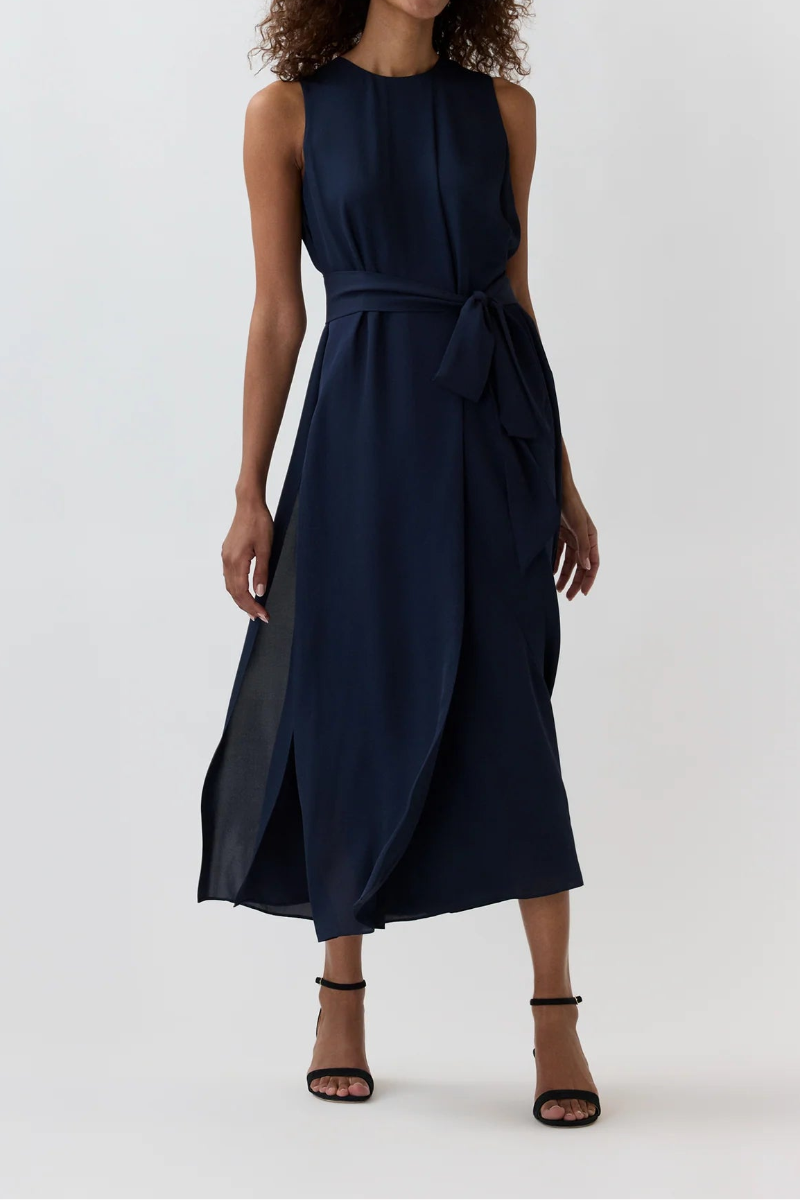 A woman modeling a sleeveless navy midi dress with a tie waist and side slit, paired with black strappy heels, against a plain light gray studio background.