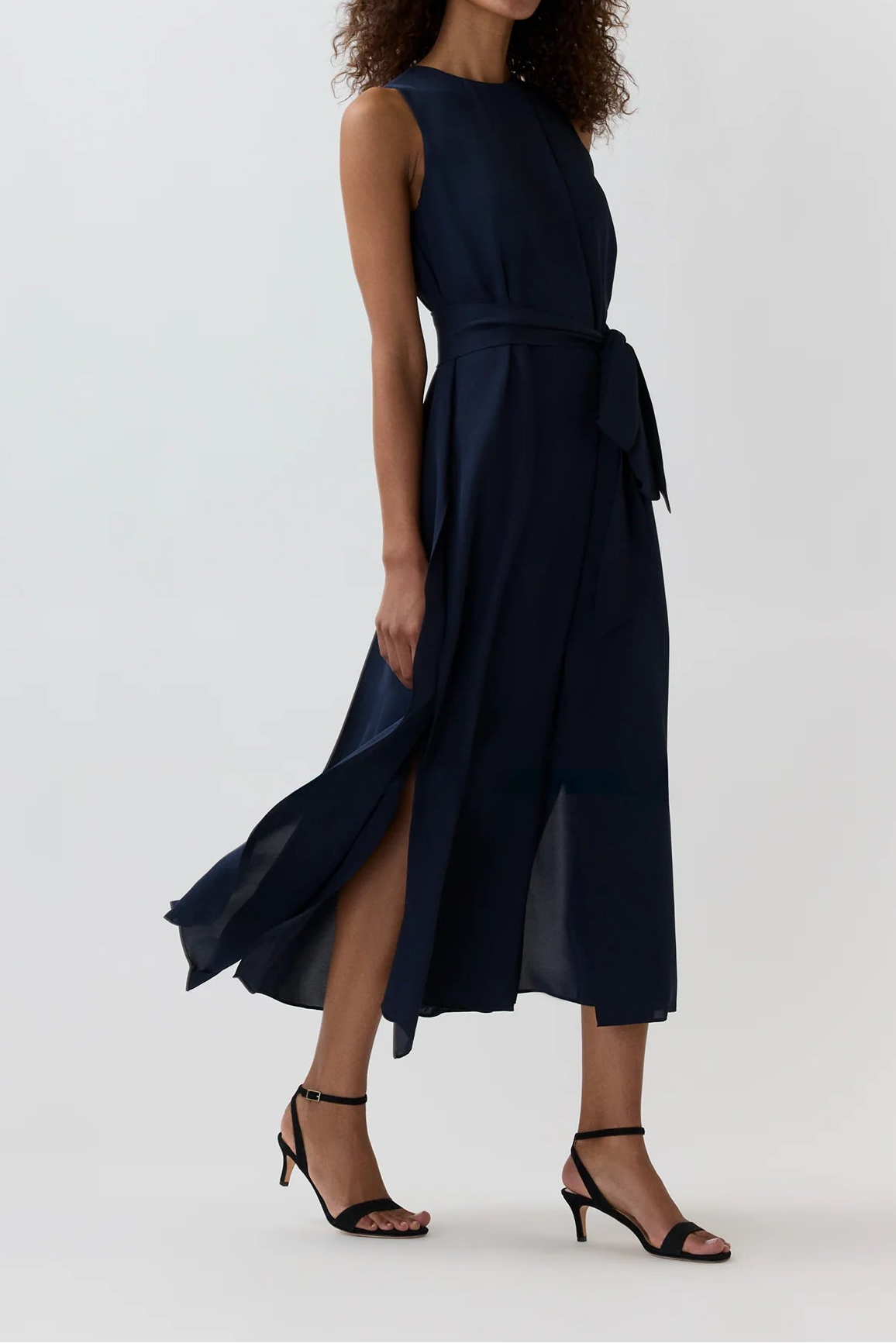 A woman modeling a sleeveless navy midi dress with a tie waist and a flowing skirt, paired with black strappy high-heeled sandals, against a plain light gray studio background.