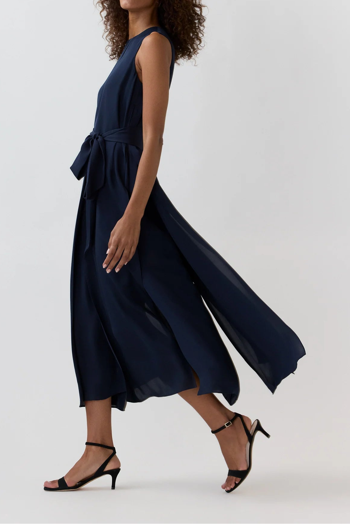A woman modeling a sleeveless navy midi dress with a tied waist and flowing skirt, paired with black strappy heels, against a light gray studio background.