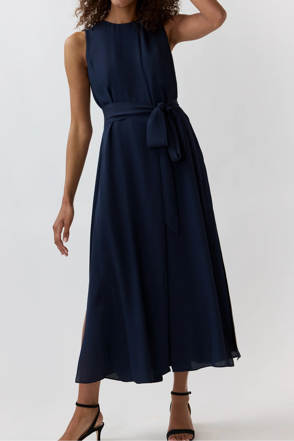 A woman modeling a sleeveless navy blue midi dress with a matching waist tie, set against a plain light gray studio background.