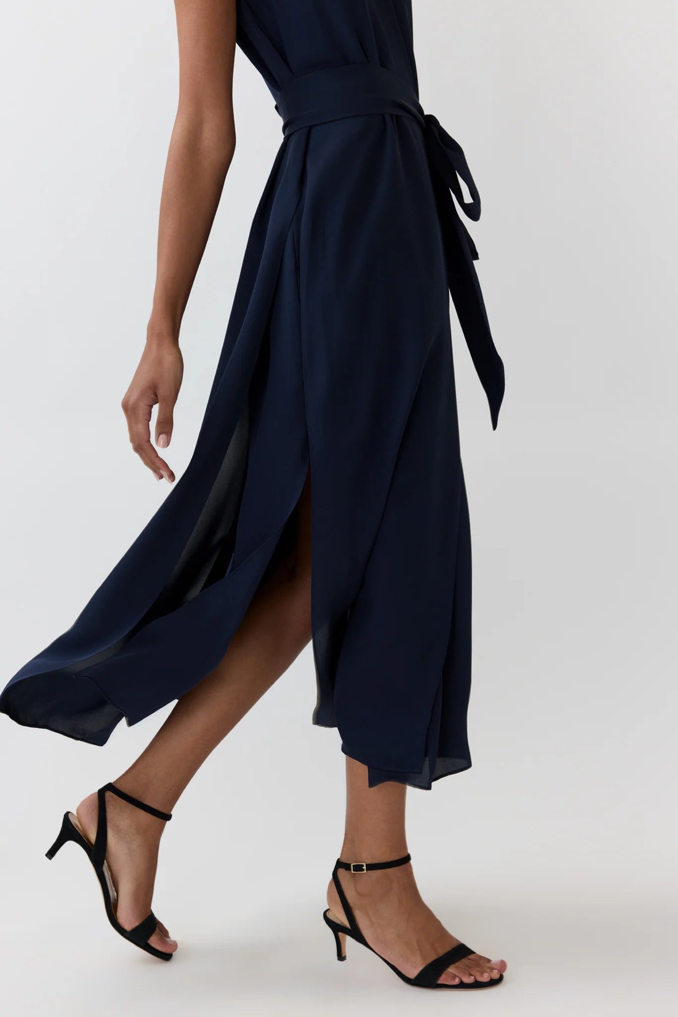 A navy blue wrap midi dress with a tie waist and side slit, shown on a model wearing black strappy high-heeled sandals against a light gray studio background.
