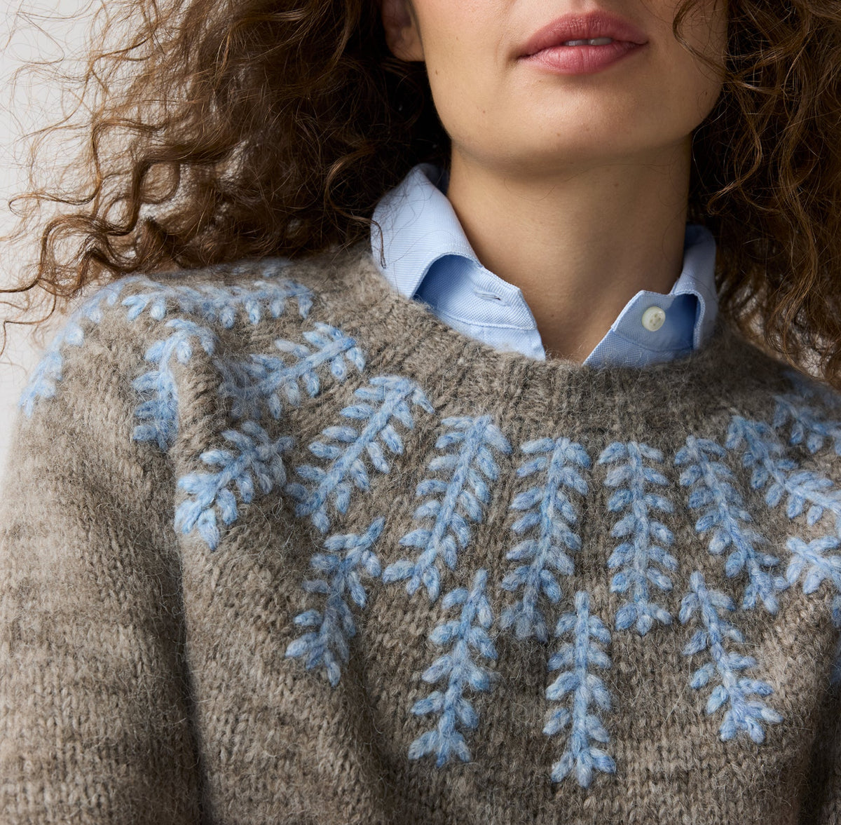 A close-up of a woman wearing a brown knit sweater with a light blue leaf pattern across the chest, layered over a light blue button-up shirt.