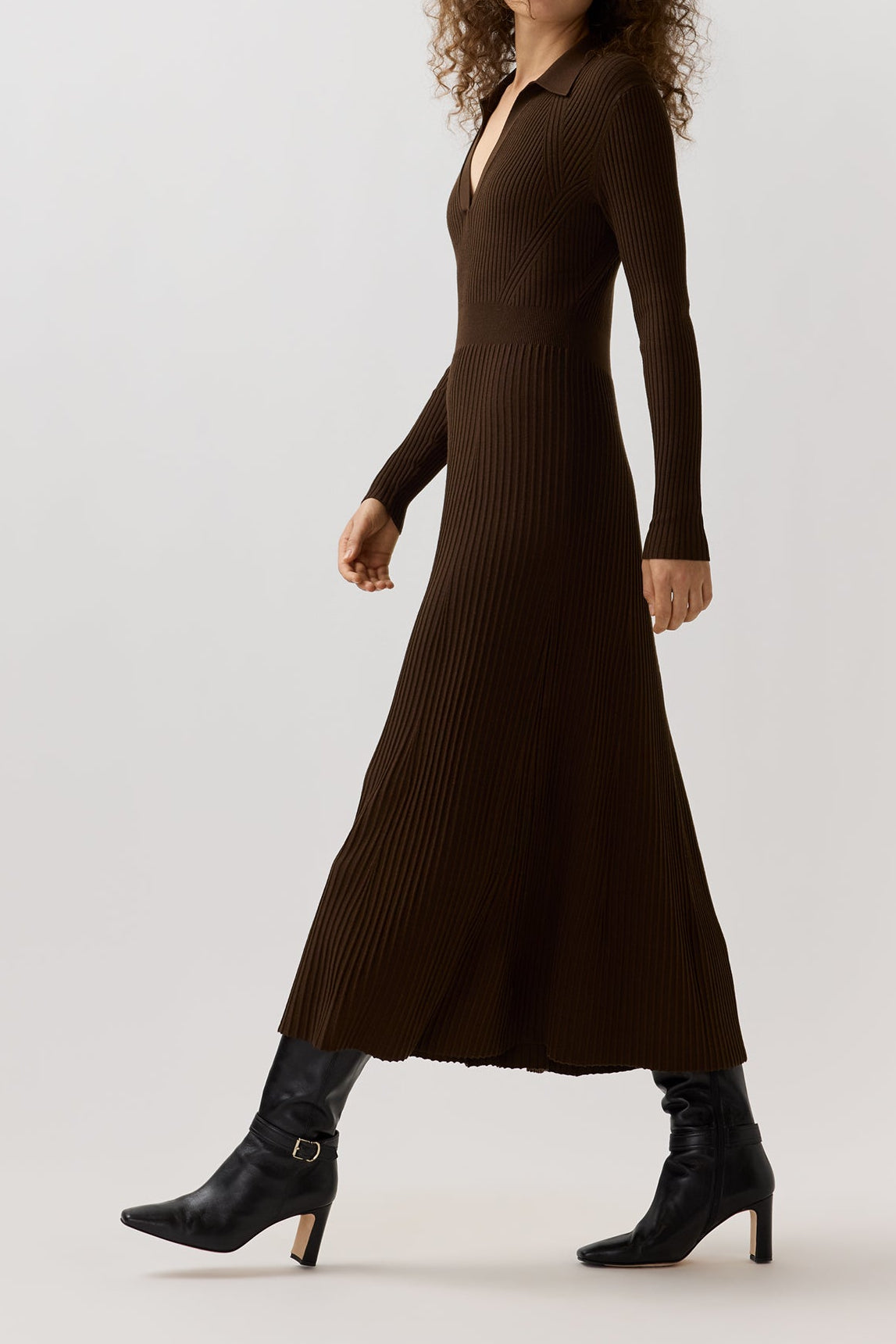 A woman modeling a dark brown ribbed long-sleeve midi dress with a V-neckline, paired with black ankle boots, against a plain light gray studio background.