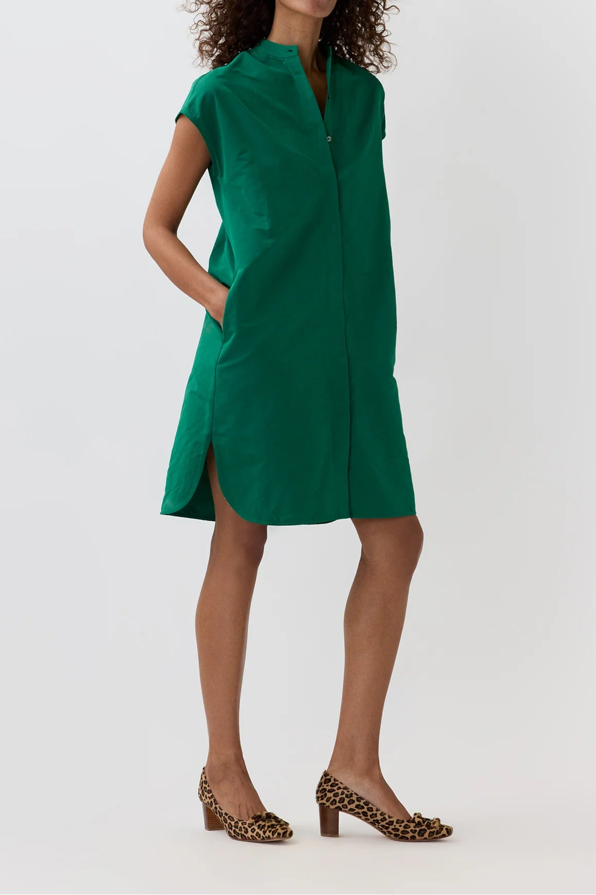 A woman modeling a green short-sleeve shirt dress with a high collar and side slits, paired with leopard-print heels, against a plain light gray studio background.