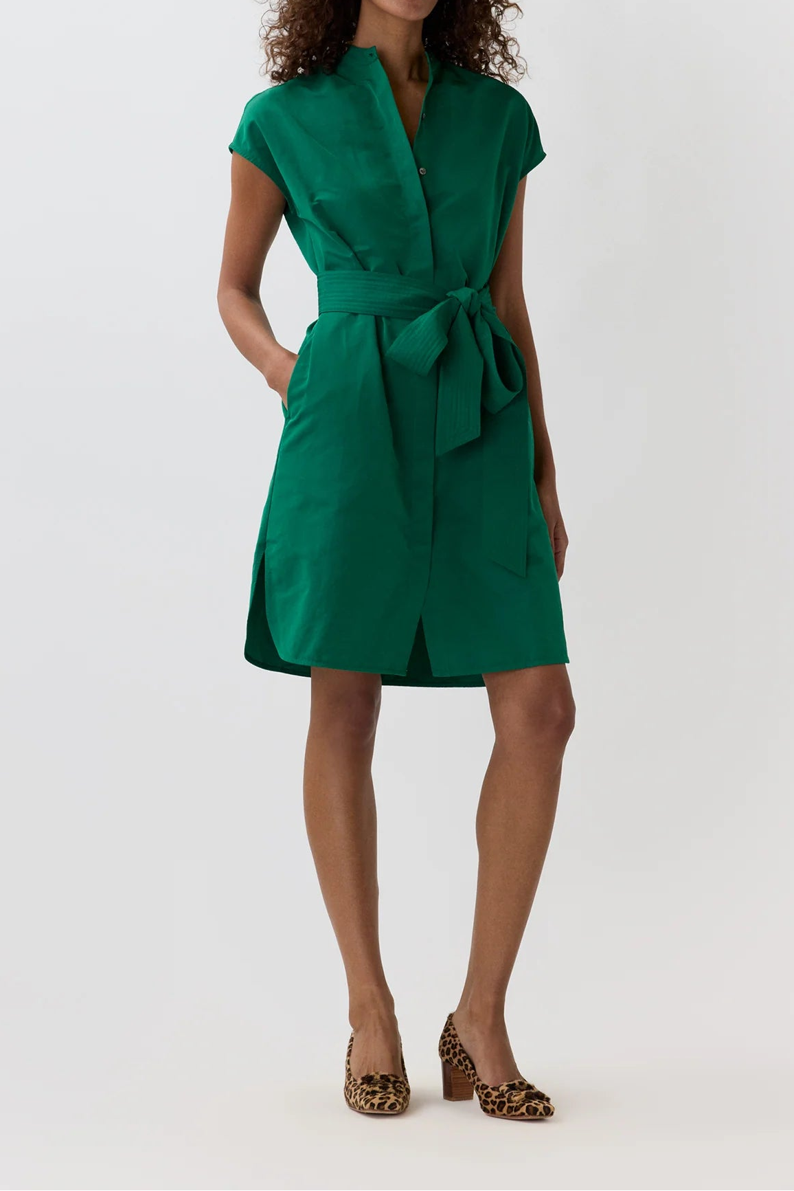 A woman modeling a short-sleeved emerald green wrap dress with a matching waist tie, paired with leopard-print heels, against a plain white studio background.
