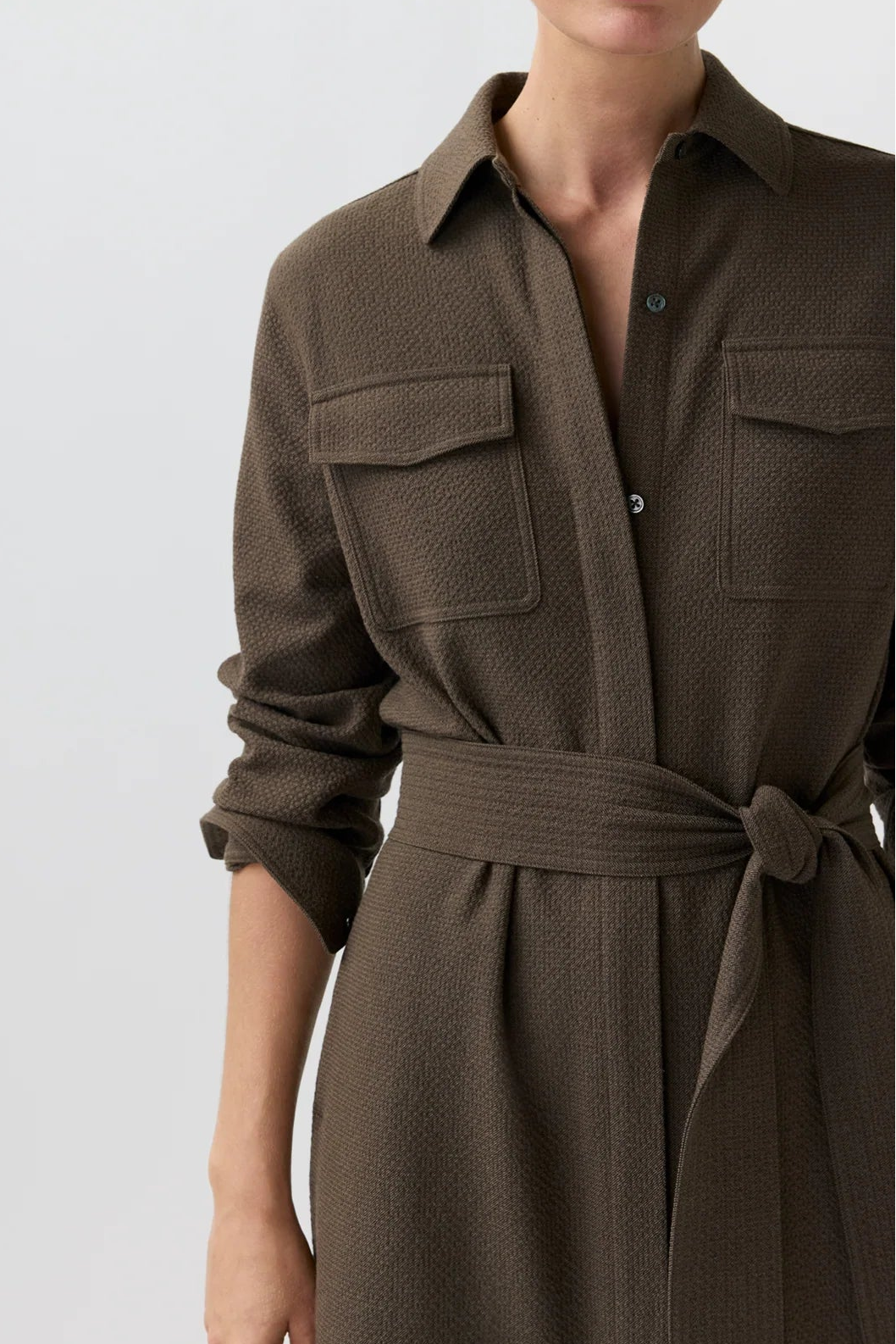 A woman wearing a dark brown textured shirt dress with a tie waist belt, shown from the chest up against a light gray studio background.